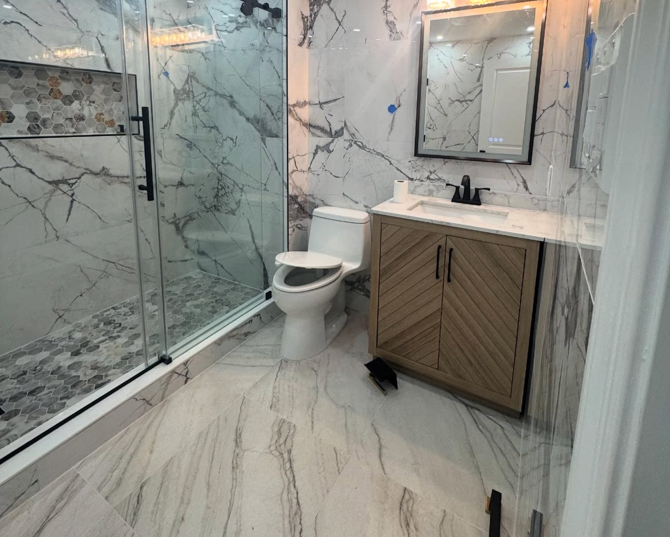 A modern bathroom with marble walls and floors, a glass-enclosed shower, a white toilet, and a wooden vanity with a white countertop and a black faucet. There is a mirror above the vanity and a roll of toilet paper on the counter.