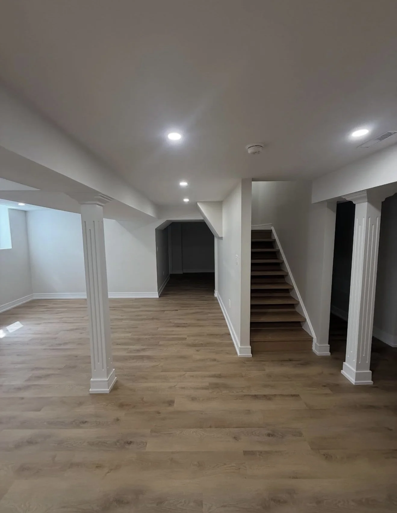 Empty room with wooden flooring, white walls, a staircase, and recessed ceiling lights.