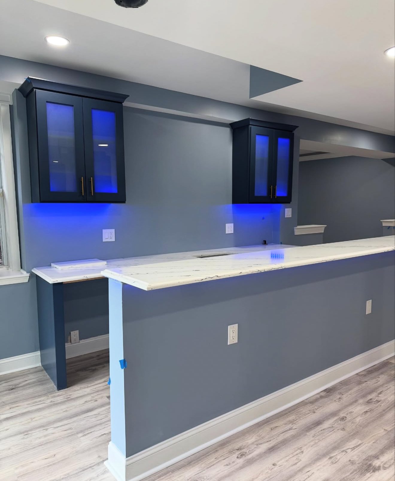 Modern kitchen island with white marble countertop, blue wall cabinets with glass doors and blue LED lighting, and gray walls with electrical outlets.