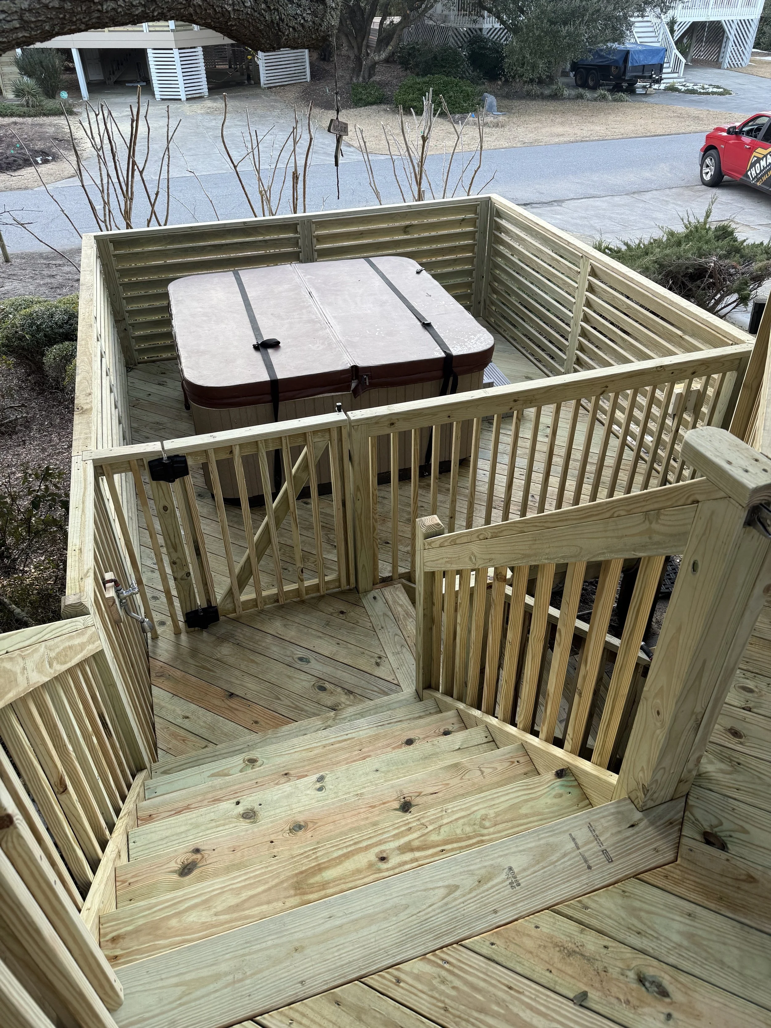 Wooden deck stairs leading down to a small enclosed deck with a hot tub, located outdoors in a residential area. The surroundings include a street, parked cars, trees, and nearby houses.