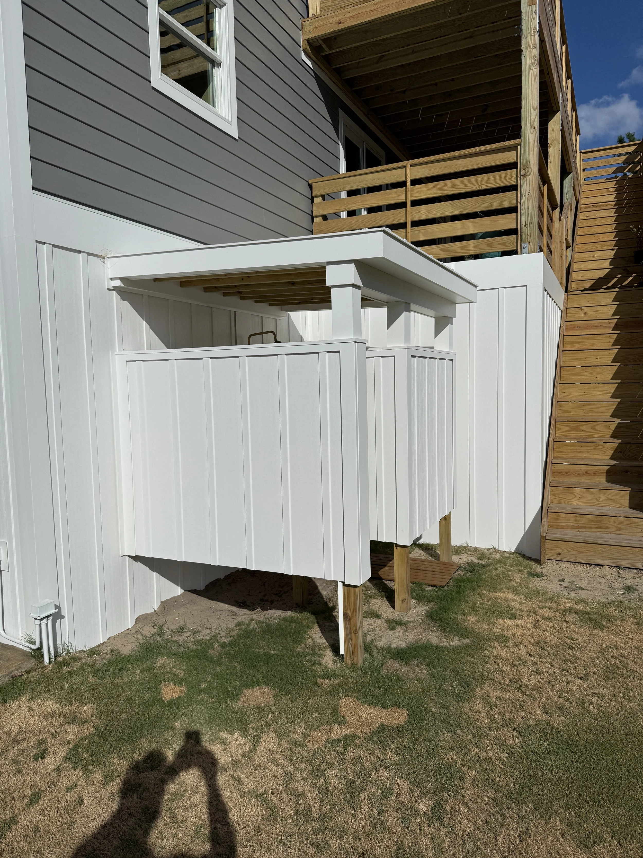 Backyard with a newly built white fence with a small gate, wooden stairs leading to a upper deck, with a two-story house with gray siding and a wooden balcony, and a clear sky.