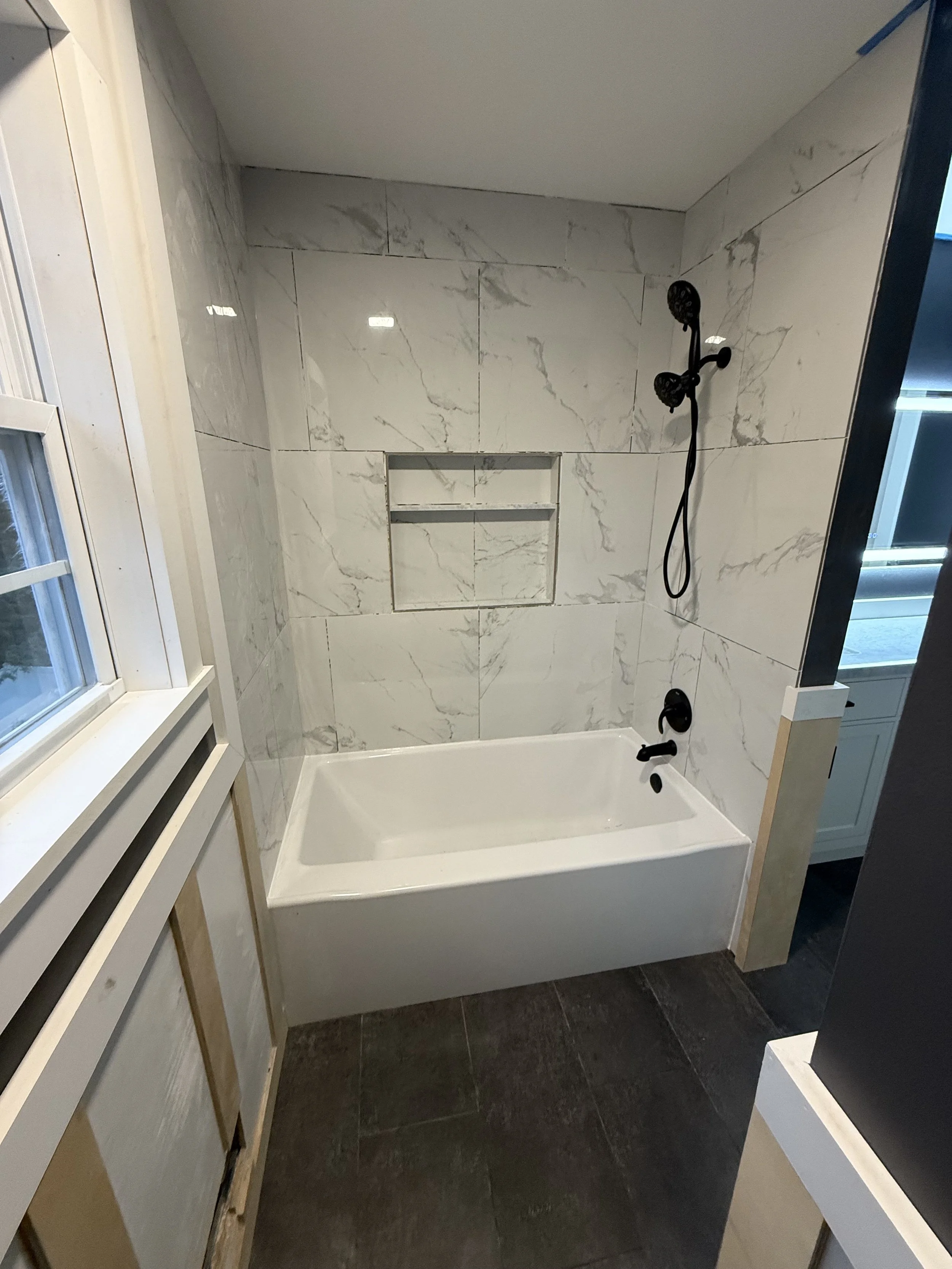 Bathroom with white marble tiled wall, built-in shelf in the wall, black showerhead and faucet, white bathtub, dark floor tiles.