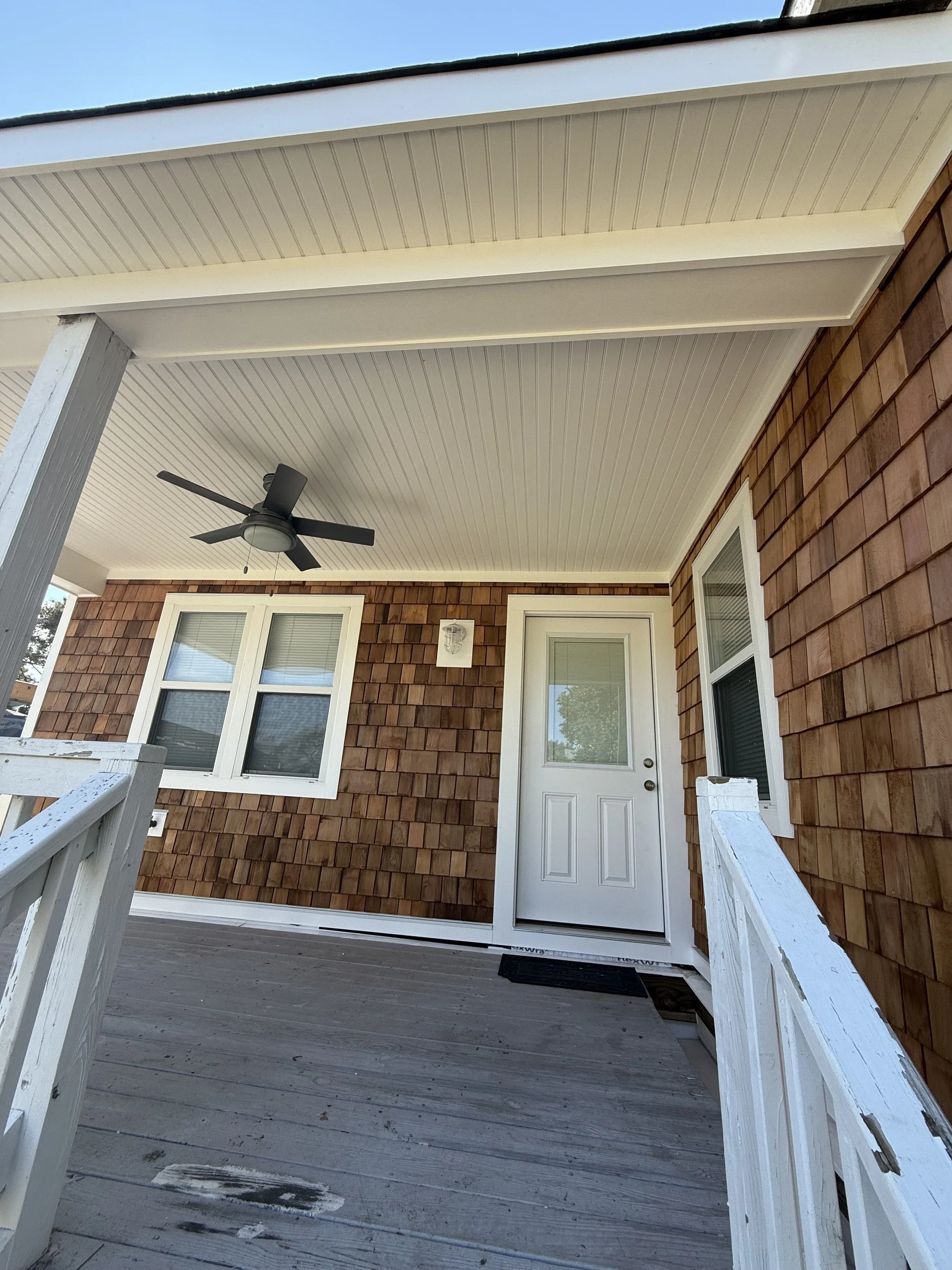 Front porch of a house with wooden flooring, white railing, a white door with a glass window, two windows, a ceiling fan, and brown shingle siding on the exterior wall.