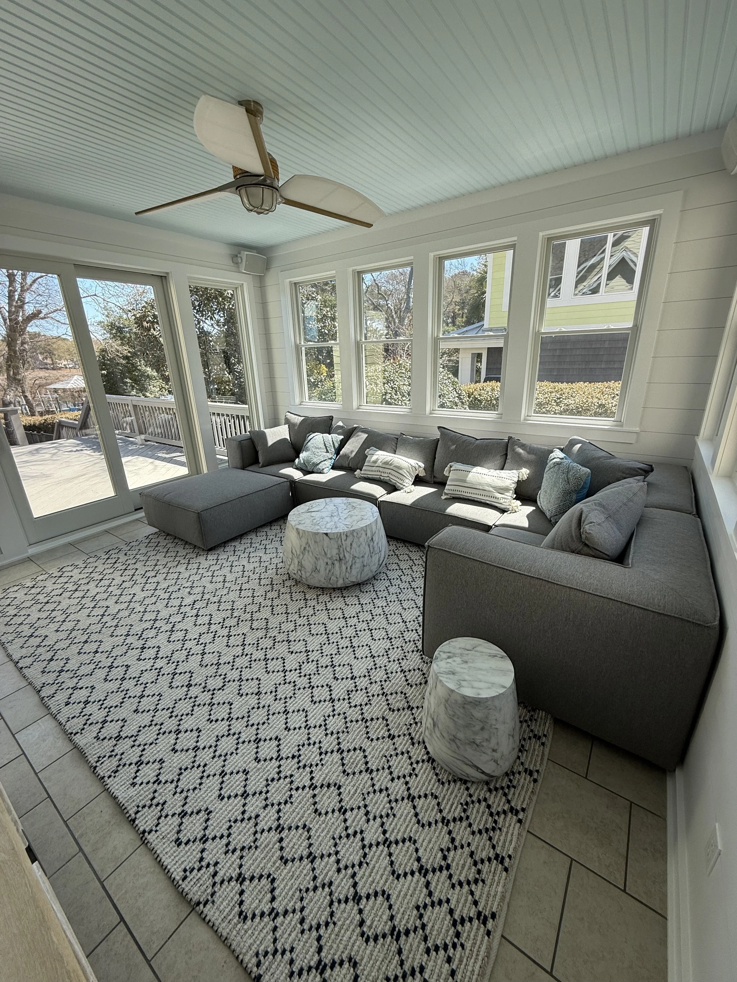Sunlit room with a gray sectional sofa, a patterned rug, two marble-topped side tables, large windows, and a ceiling fan with leaf-shaped blades.