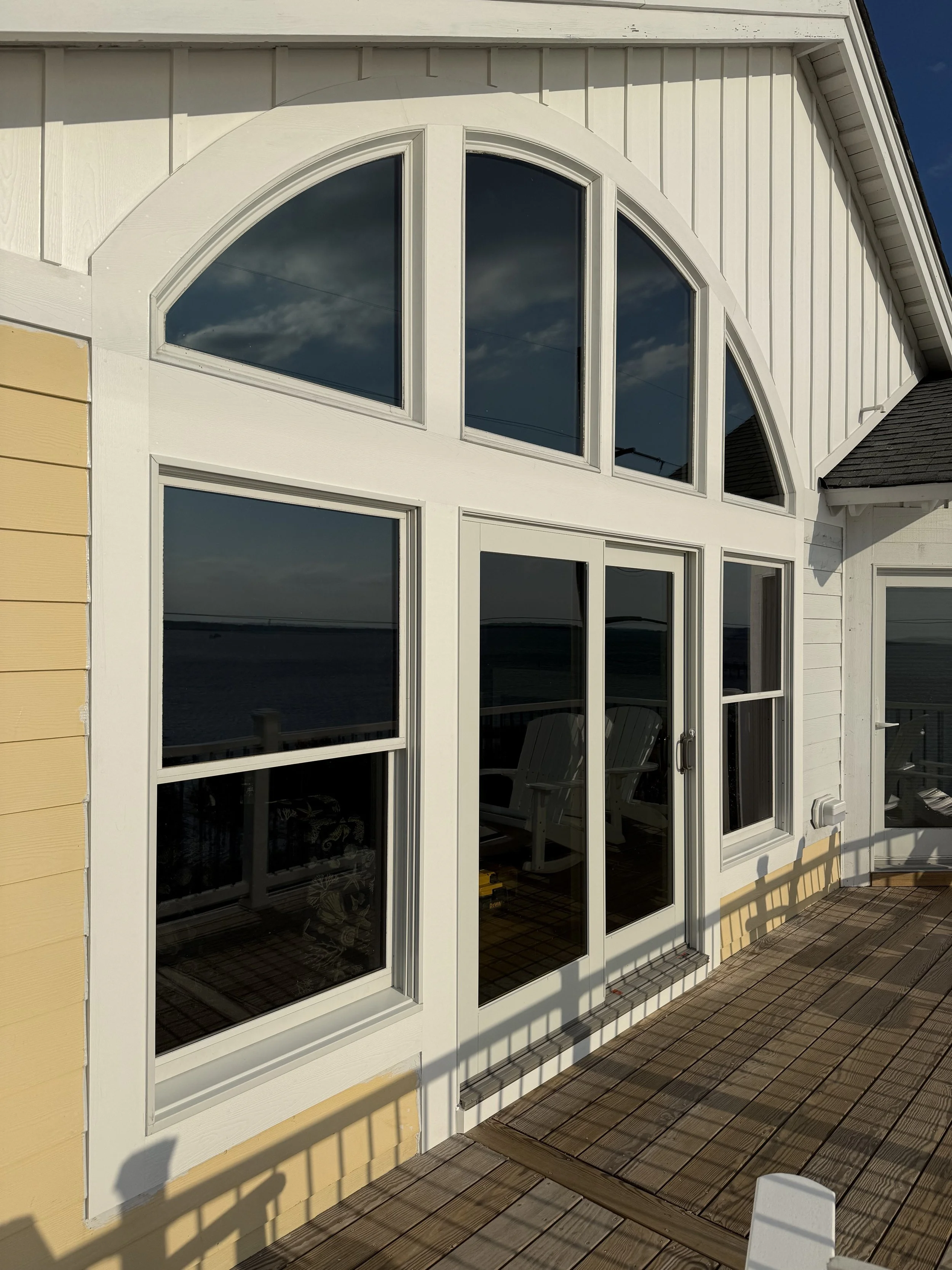 A house with large white-framed glass windows and door on a wooden deck, overlooking water with puffy clouds in the sky.