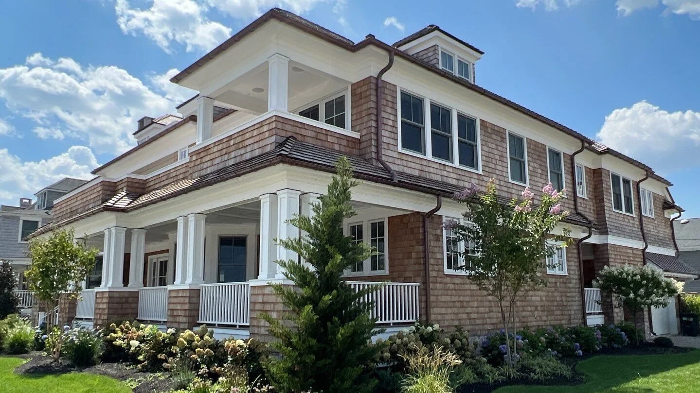 A large, multi-story house with a brick exterior, white columns, and a porch, surrounded by well-maintained landscaping and flowering trees under a partly cloudy sky.