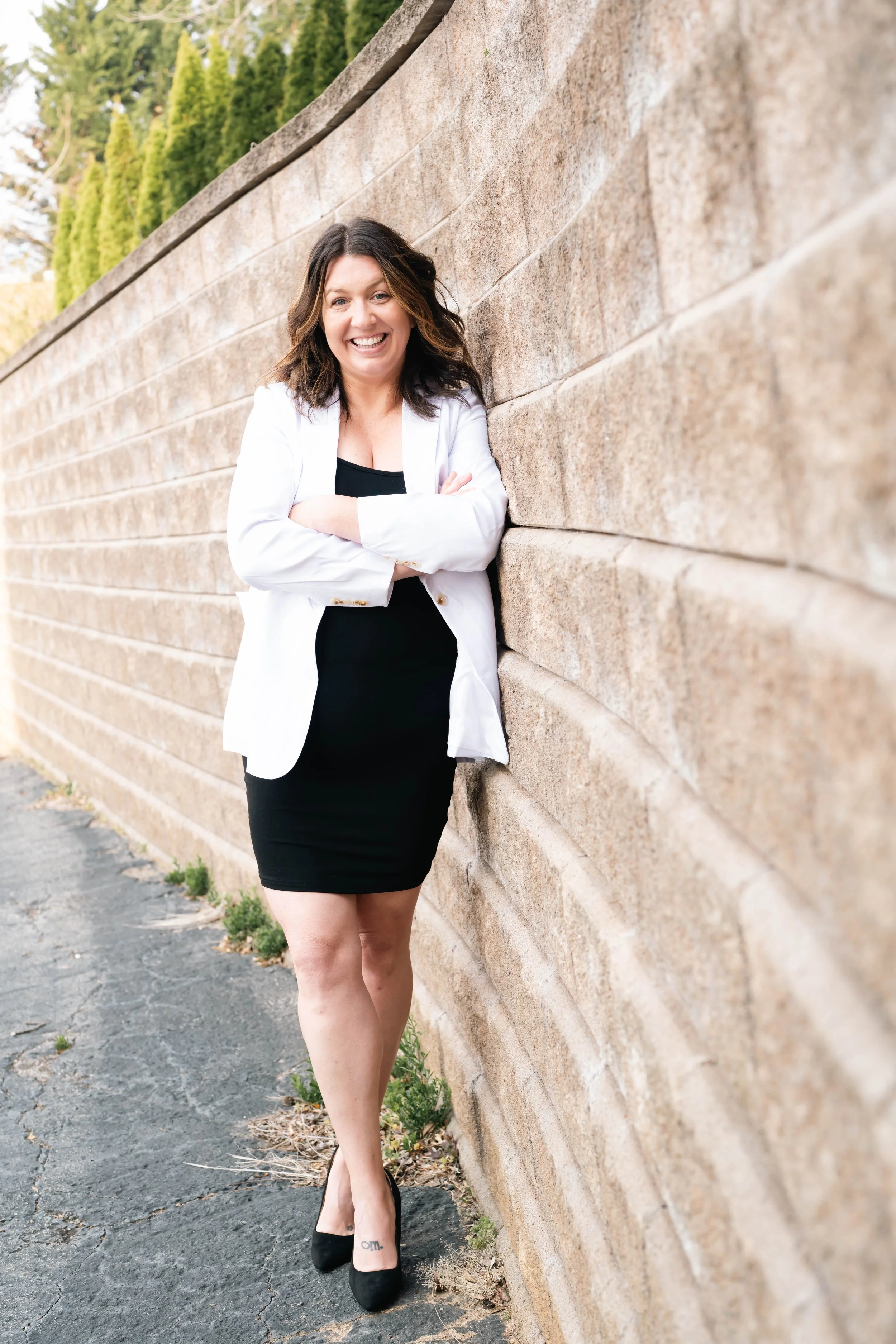 A woman smiling, wearing a black dress and white blazer, standing outdoors against a stone wall with green trees in the background.