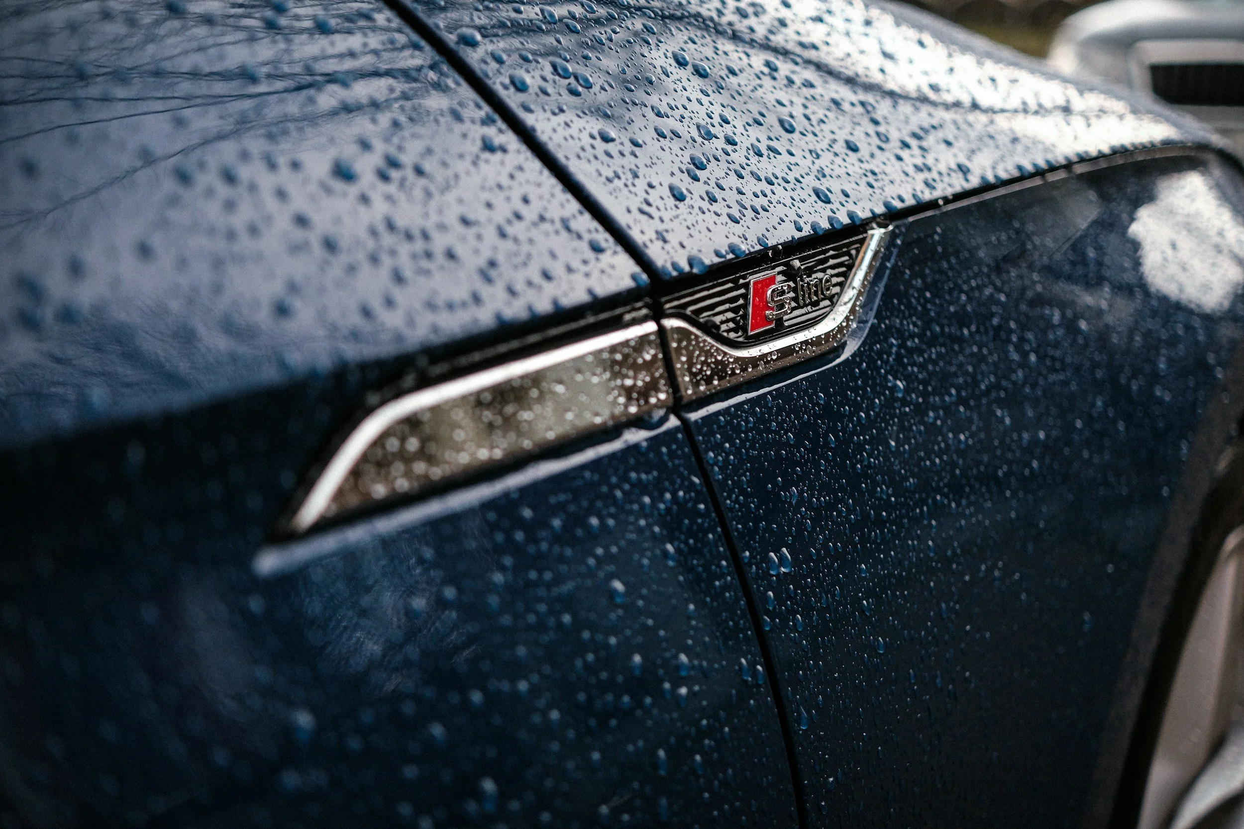 Close-up of a blue car with raindrops on the surface, showcasing a badge that reads "S line".