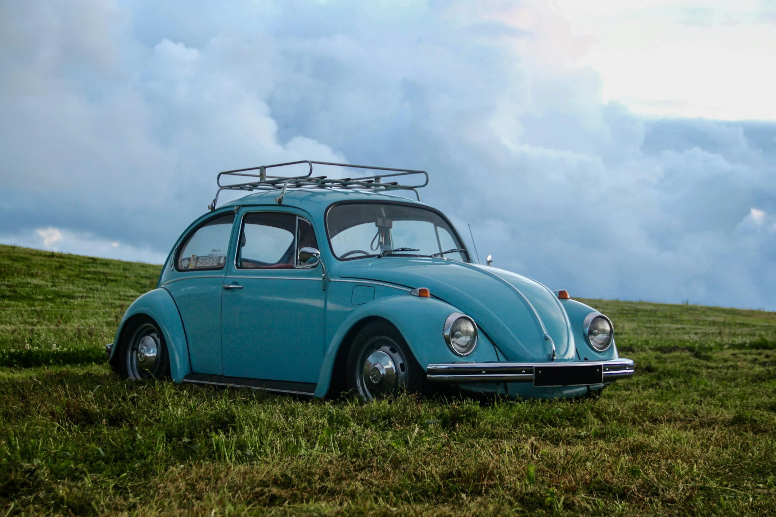 A vintage blue Volkswagen Beetle with a roof rack parked on a grassy field under a cloudy sky.