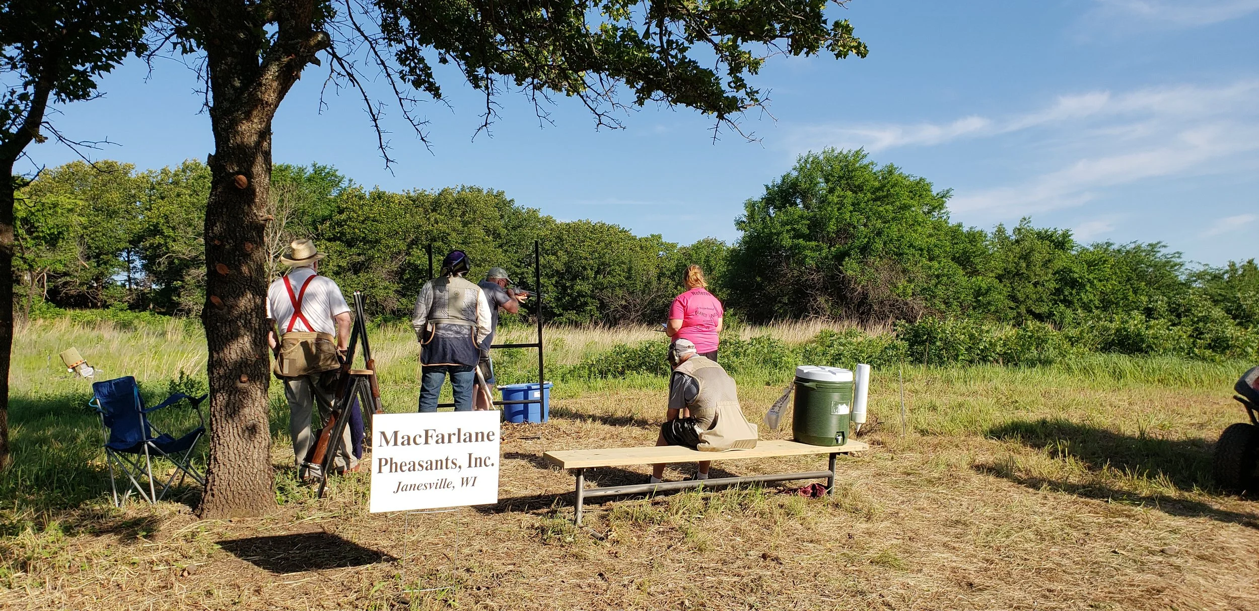 People participating in a shooting event outdoors, with targets and equipment set up, and a sign that reads 'MacFarlane Pheasants, Inc. Janesville, WI'.