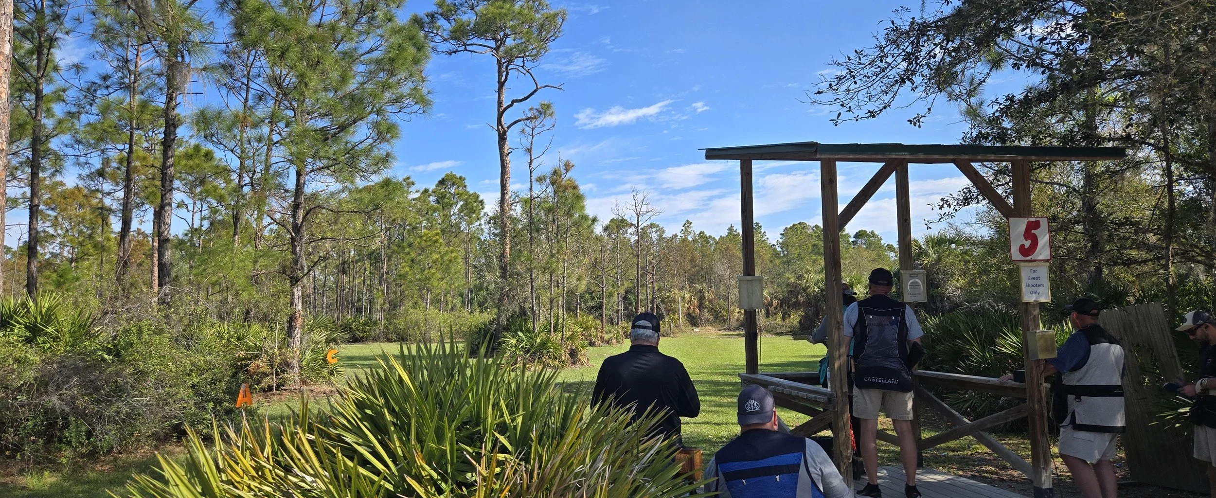 People playing disc golf on a course with trees and greenery, with a wooden tee area and a sign that says '5' and 'For Event Shooters Only'.