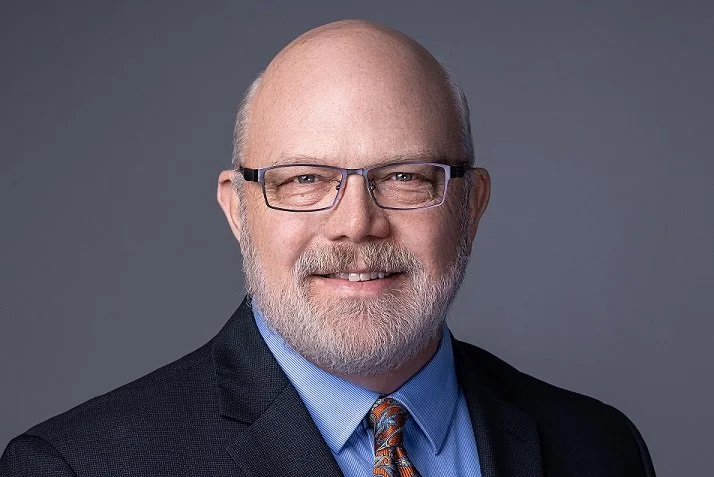 Portrait of a middle-aged man with glasses, a beard, and a bald head, wearing a dark suit, light blue shirt, and patterned tie, smiling against a gray background.
