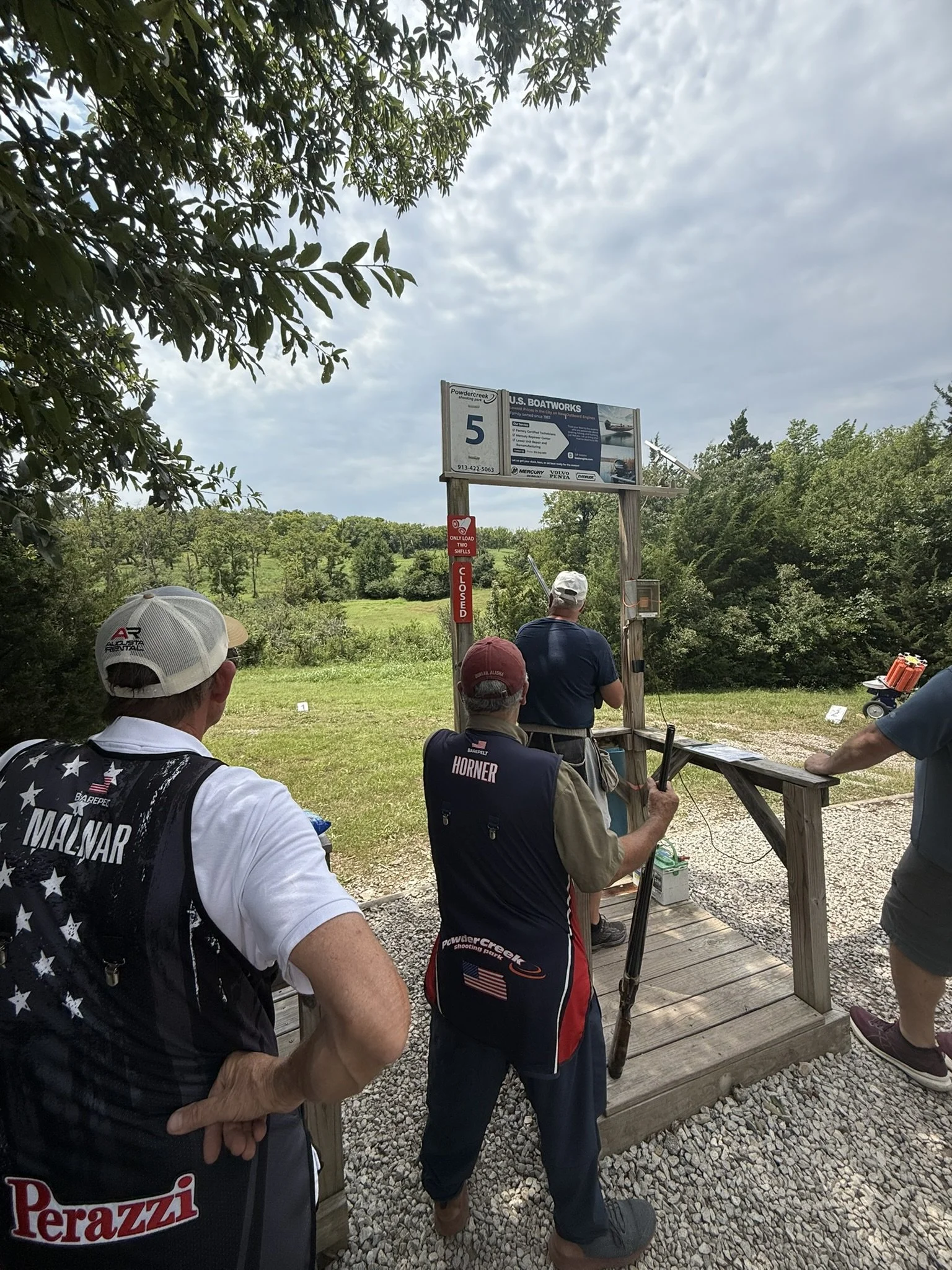 Group of people at a shooting range, aiming at targets, with trees and cloudy sky in background.