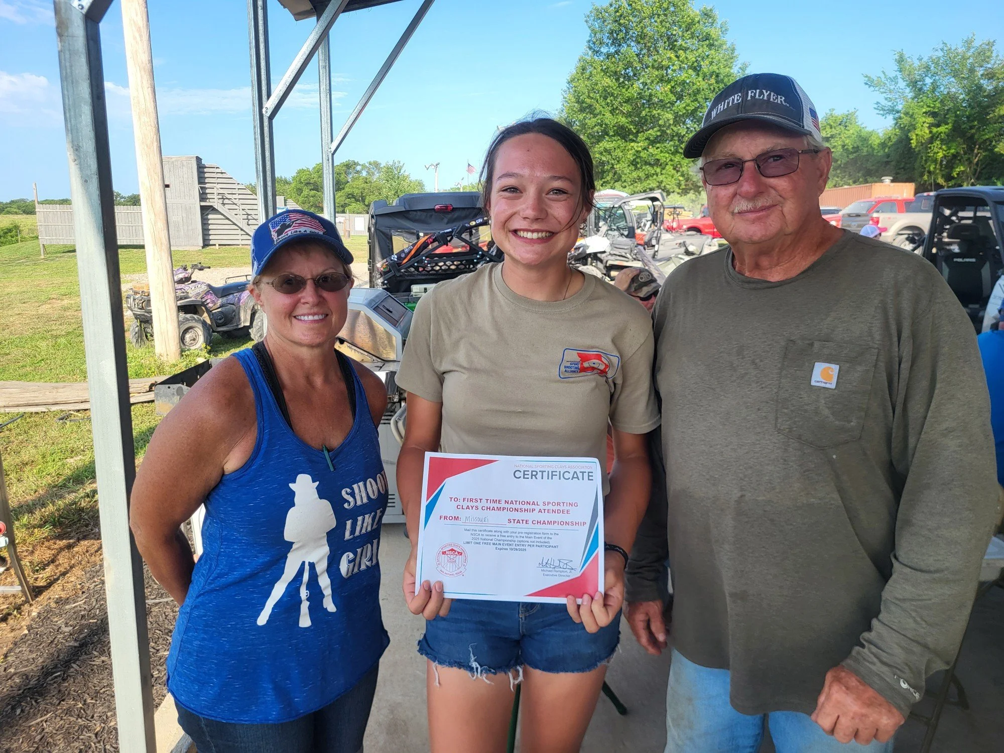 Three people standing outdoors with a young girl holding a certificate, smiling at the camera. Two adults, a woman in a blue tank top and a man in a gray shirt, stand on either side of the girl. The background shows green trees, outdoor equipment, an