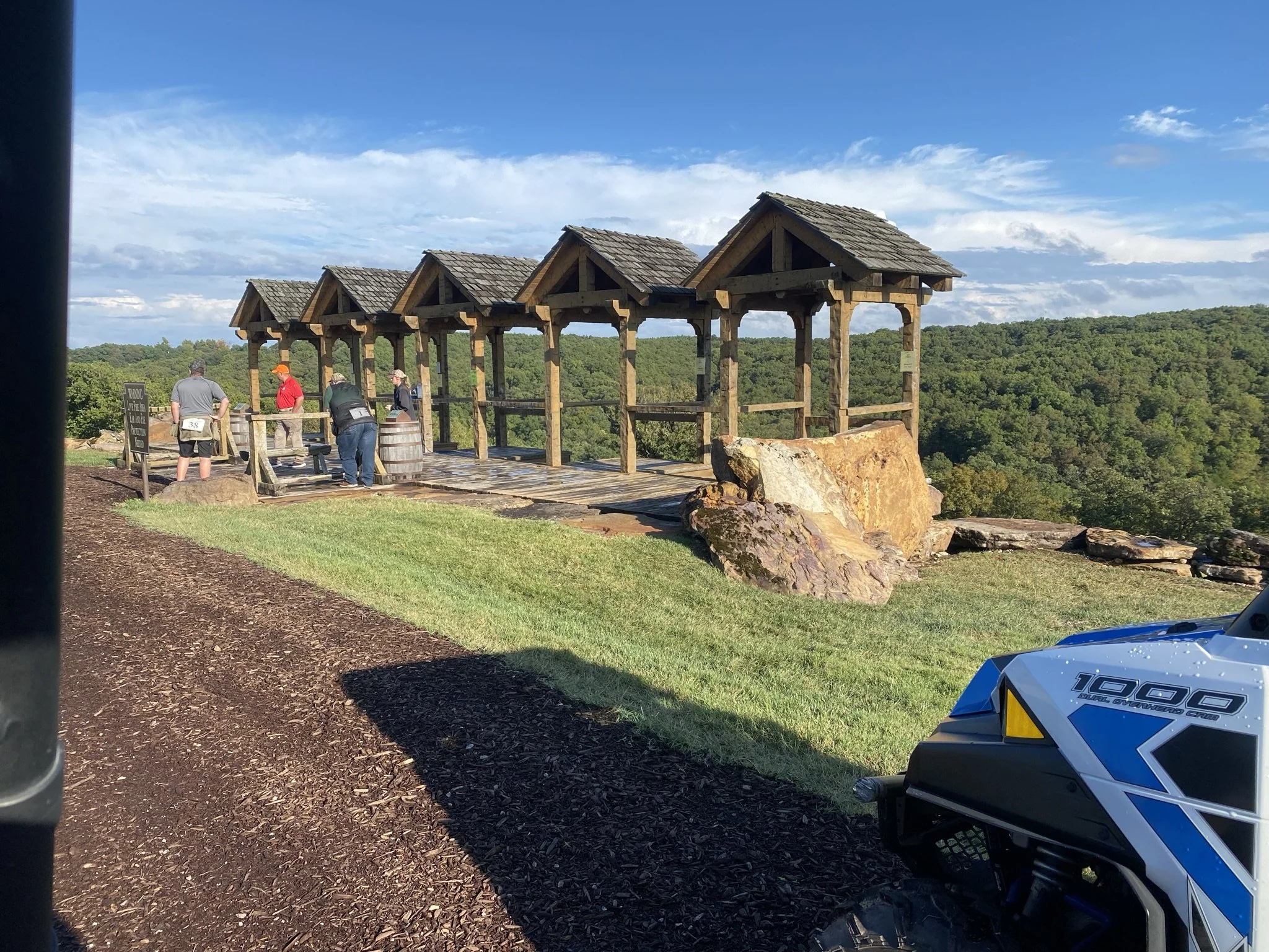 A scenic view of a wooden overlook with four covered sections and a large rock in front, overlooking a green landscape with trees under a partly cloudy sky. Several people are gathered at the overlook. Part of a blue and white all-terrain vehicle is 
