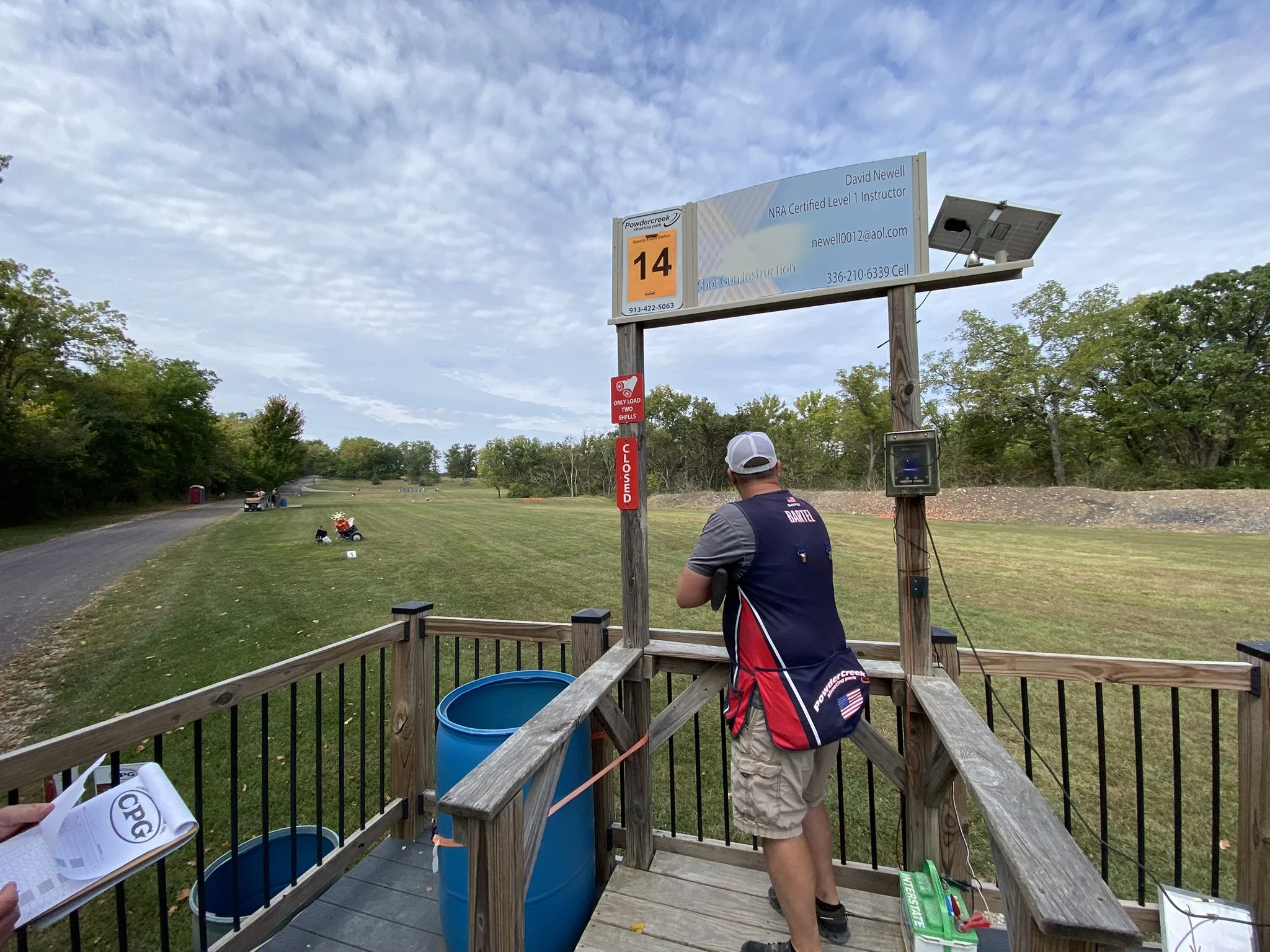 A man with a gray cap and a navy and red shooting vest stands on a wooden platform at a shooting range, with a gun in his hands. There are two blue trash cans next to him, and a person holding a pamphlet or score sheet in the foreground. The range is