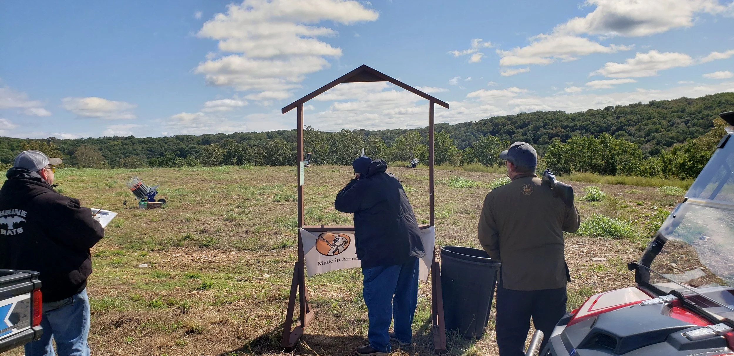 People participating in a shooting practice or competition outdoors on a sunny day, with targets set up in the distance and a wooded hill in the background.