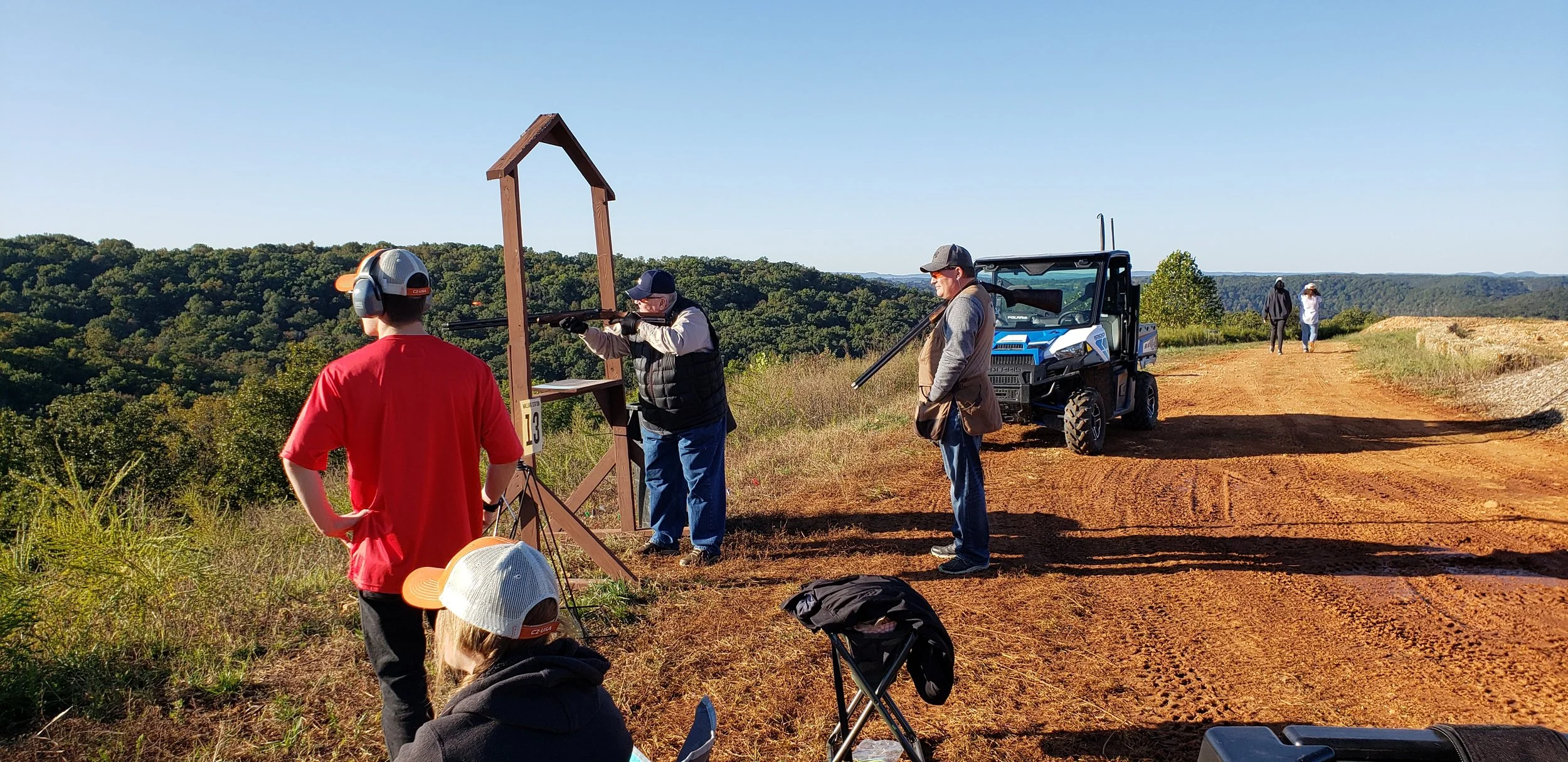 People participating in a clay pigeon shooting event outdoors on a sunny day, with some aiming shotguns at targets, others observing, a utility vehicle on a dirt path, and trees and hills in the background.