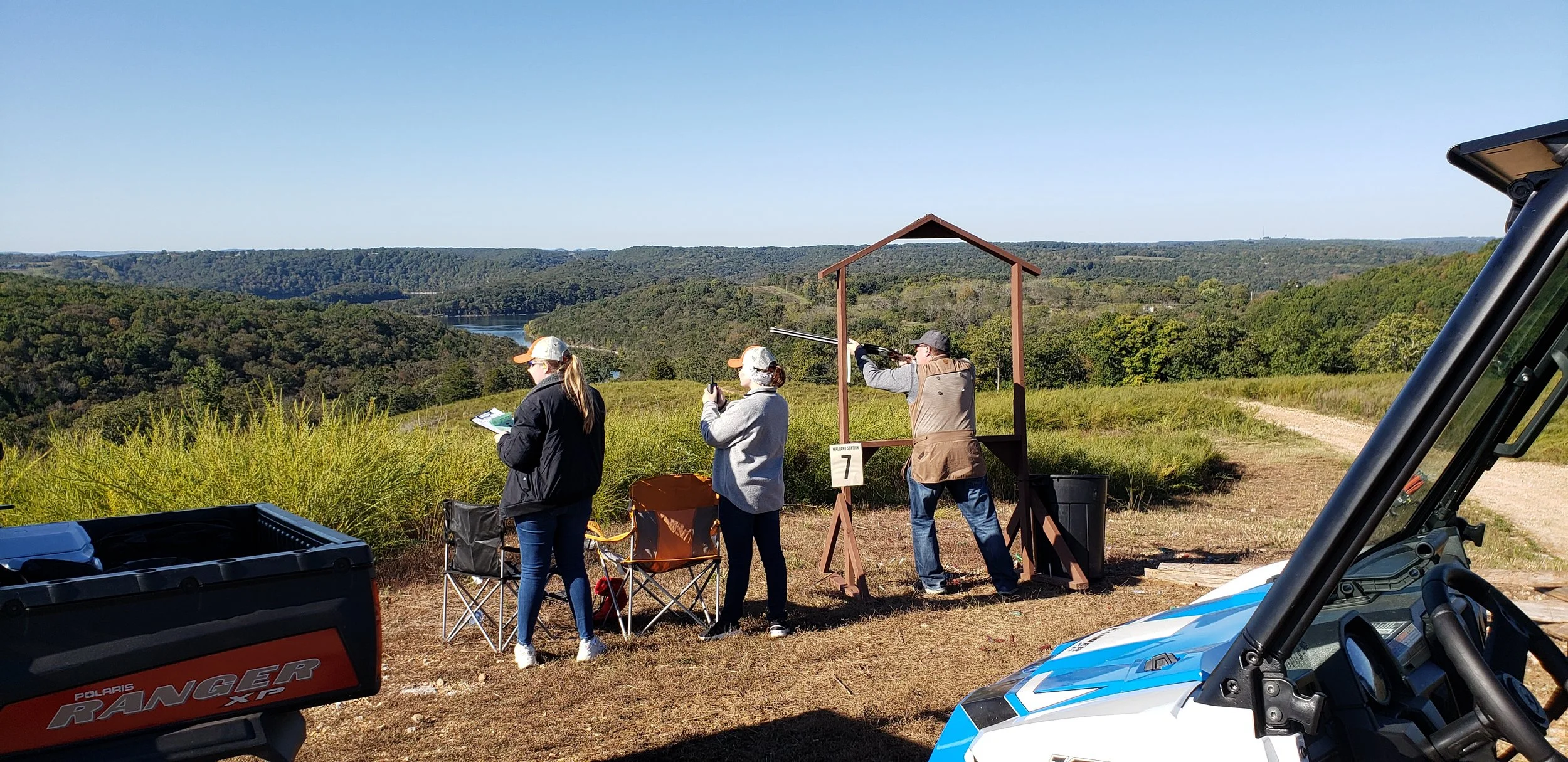 People engage in a shooting activity outdoors on a clear day, with hills and a river in the background. One person is aiming a rifle within a wooden shooting structure, while others observe or prepare, near a Polaris Ranger XP vehicle.
