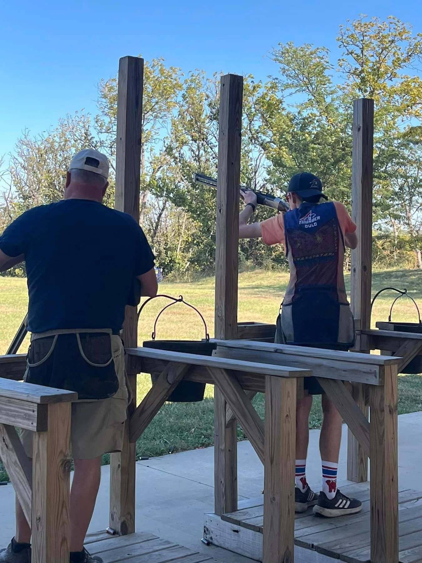 A young boy shooting a shotgun at a firearm range, overseen by an adult man.