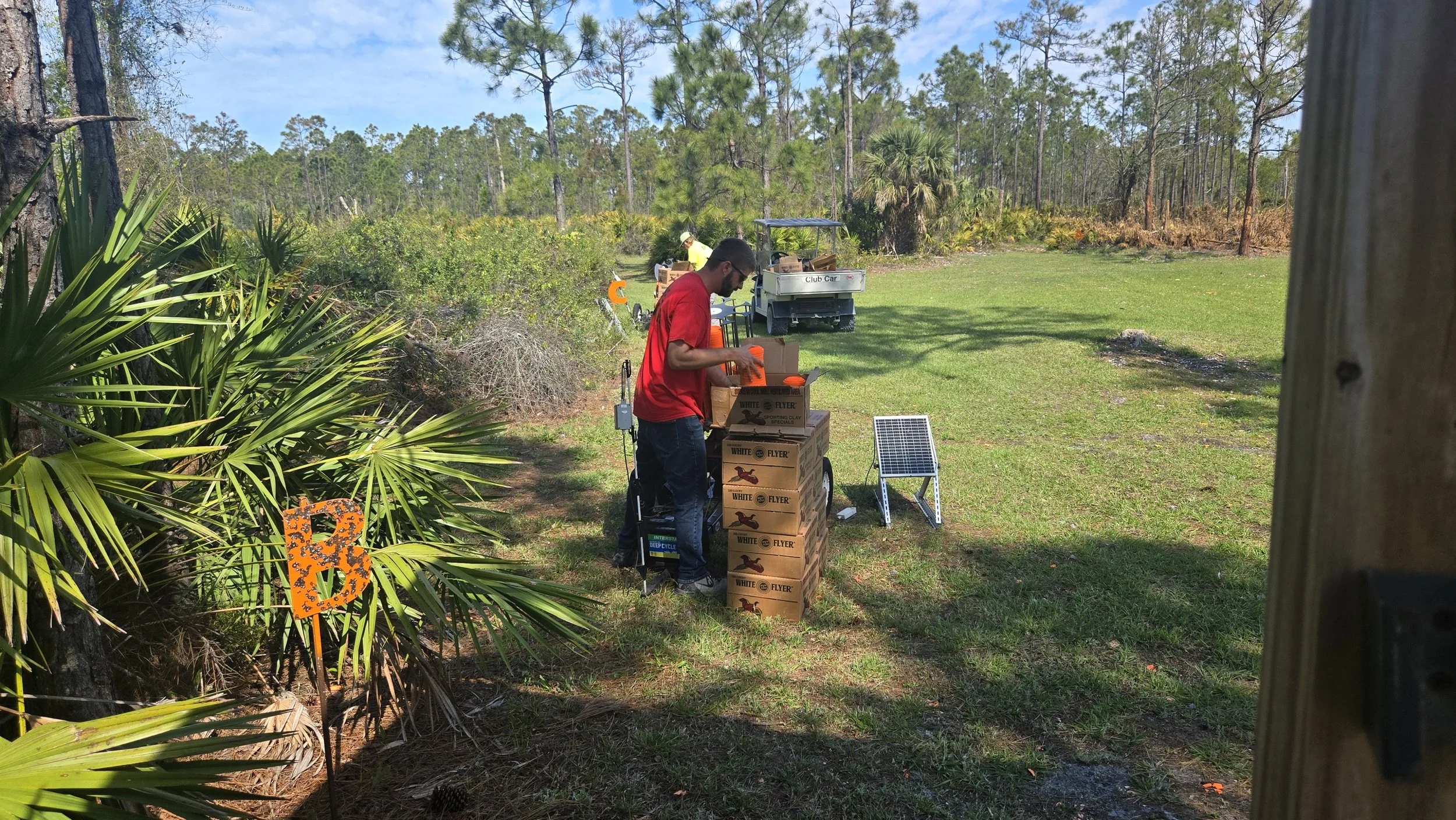 Two men preparing golf course maintenance supplies near a golf cart on a sunny day. One man is stacking boxes labeled 'White Flyer,' and the other man is in the background near a small utility vehicle. The scene is in a lush, green outdoor setting wi