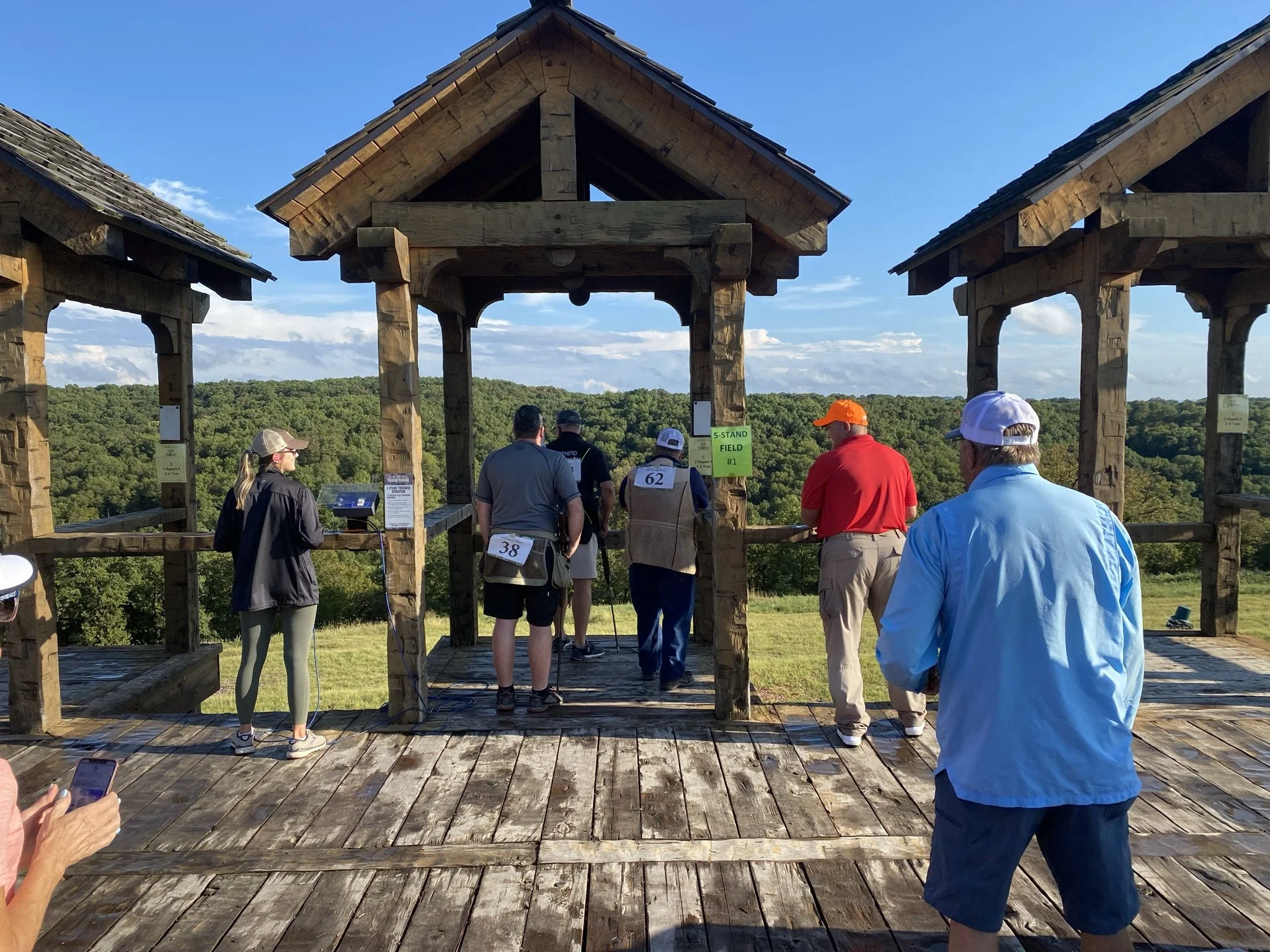 A group of people standing on a wooden platform outdoors, preparing to hit golf balls from an elevated tee area with a scenic view of trees and a blue sky.