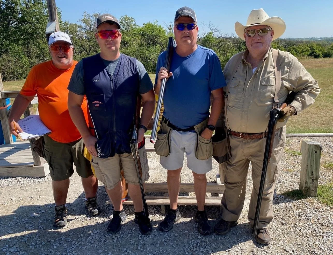 Four men standing outdoors, dressed in casual outdoor clothing with three holding shotguns, on a gravel surface with greenery background.
