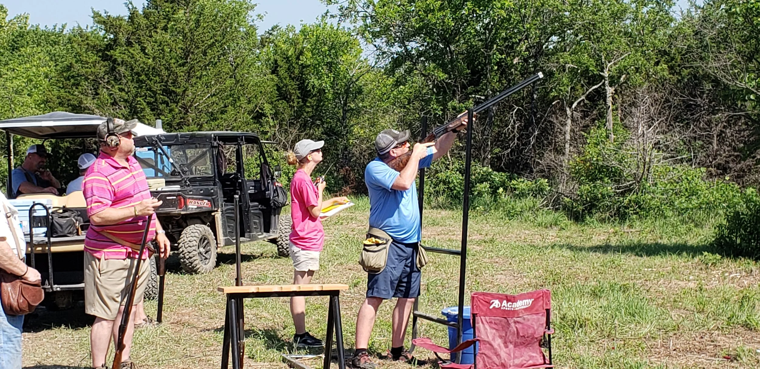 People participating in an outdoor shooting event. One person is aiming a shotgun at a target, while others observe. The scene takes place in a grassy field with trees in the background.