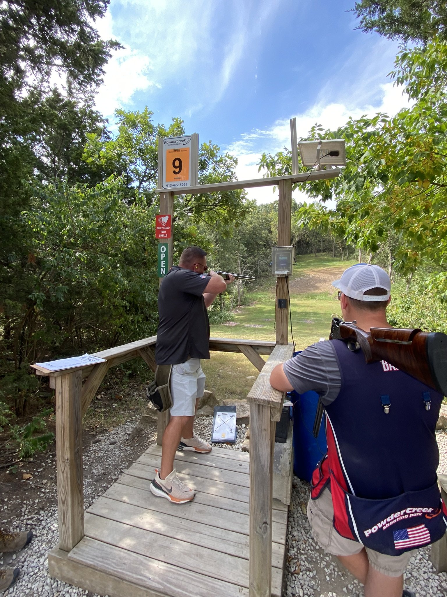 Two men at a shooting range, one aiming a rifle and the other standing nearby with a shotgun on his shoulder, surrounded by trees and a blue sky.