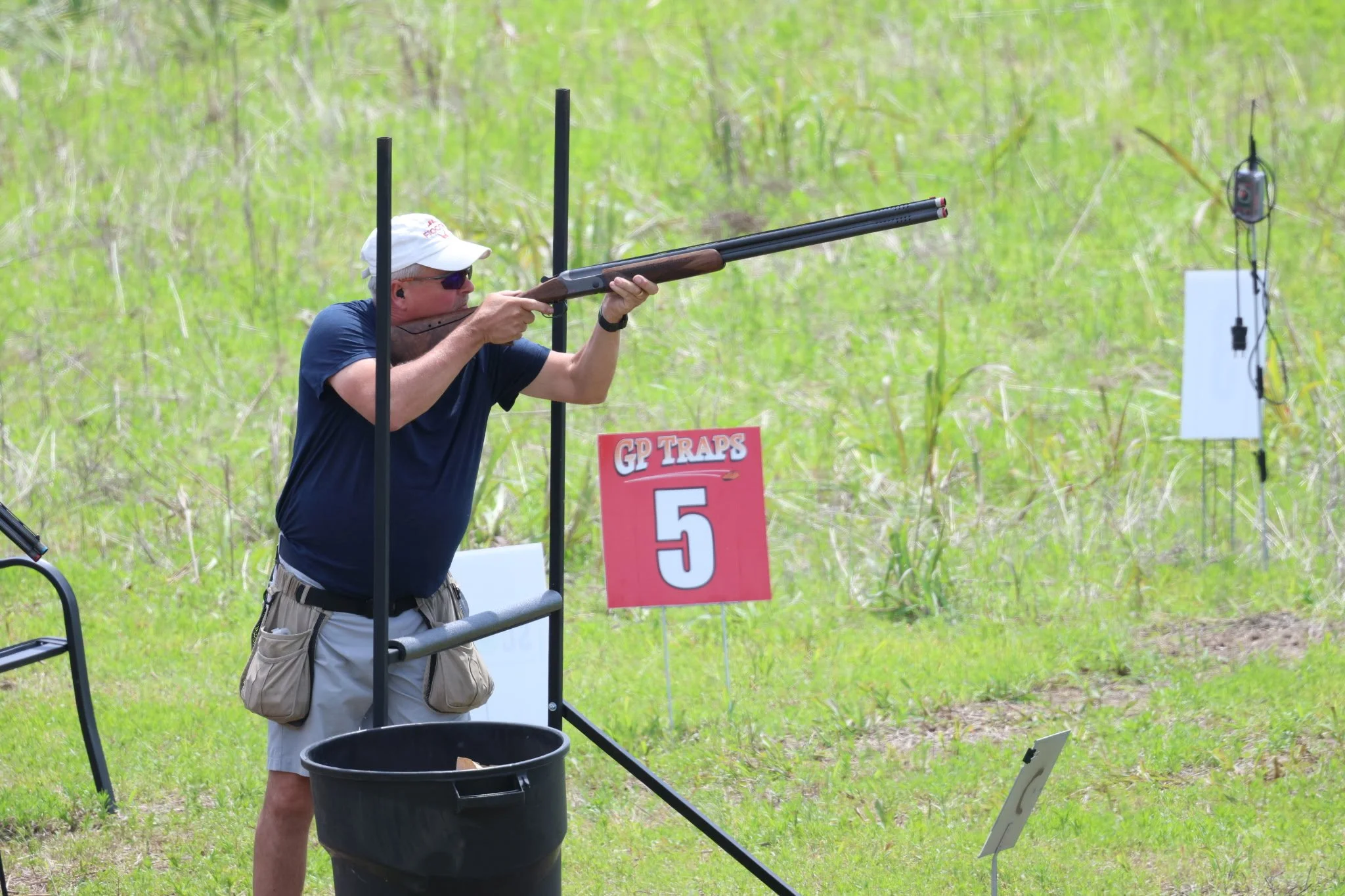 A man aiming a shotgun at a shooting range, with a sign reading 'GP Traps 5' and a target in the background, outdoors in a grassy field.