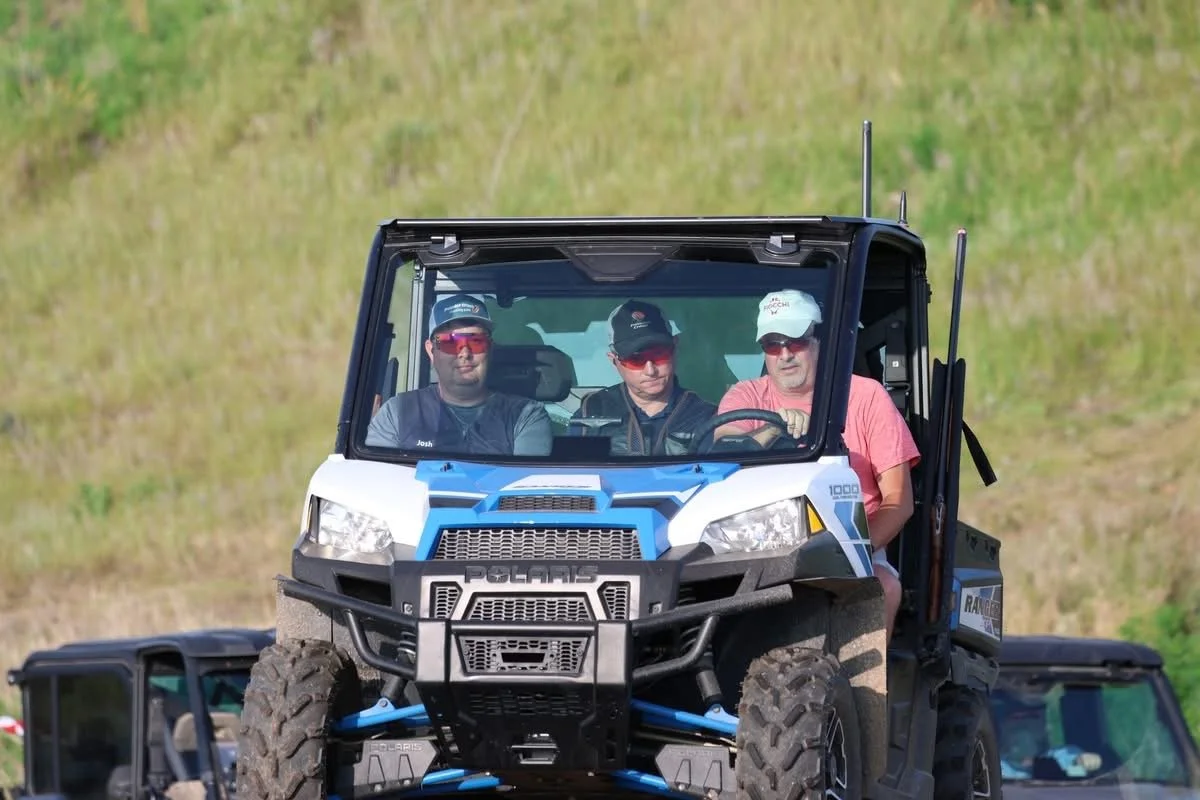 Three men sitting inside a Polaris off-road utility vehicle, on a grassy outdoor terrain.