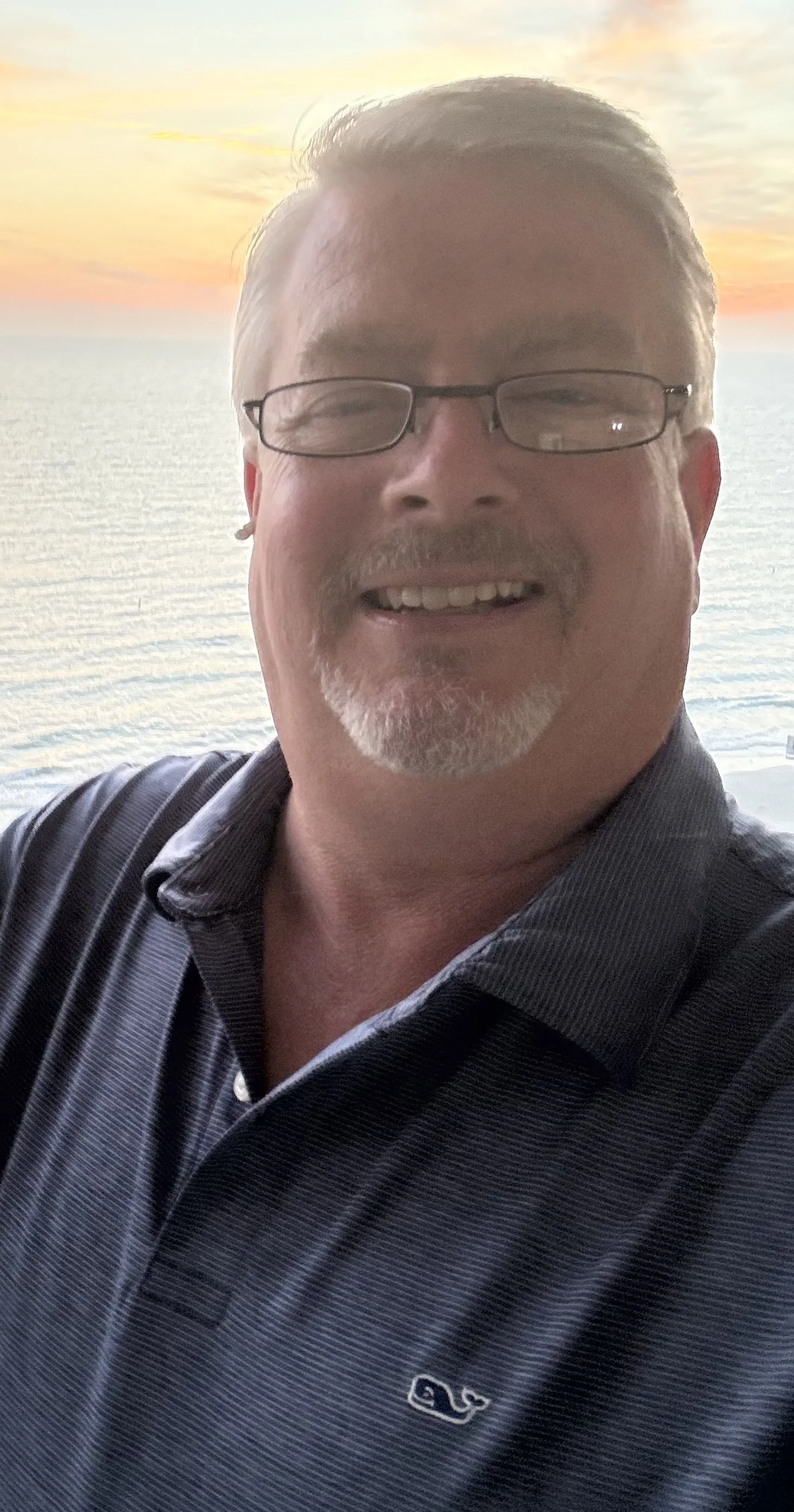 A man smiling for a selfie with ocean and sunset in the background.