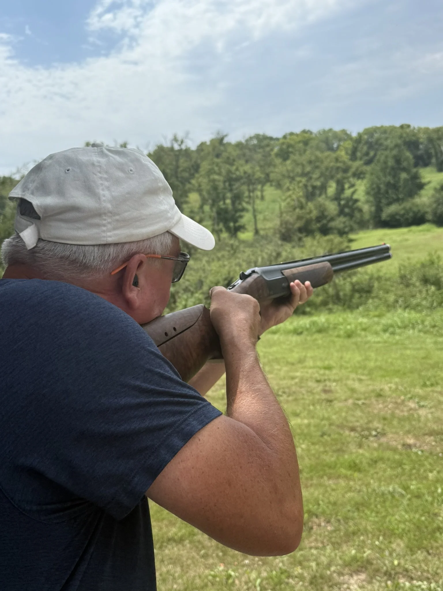 An older man aiming a shotgun outdoors on a grassy field with trees and blue sky in the background.