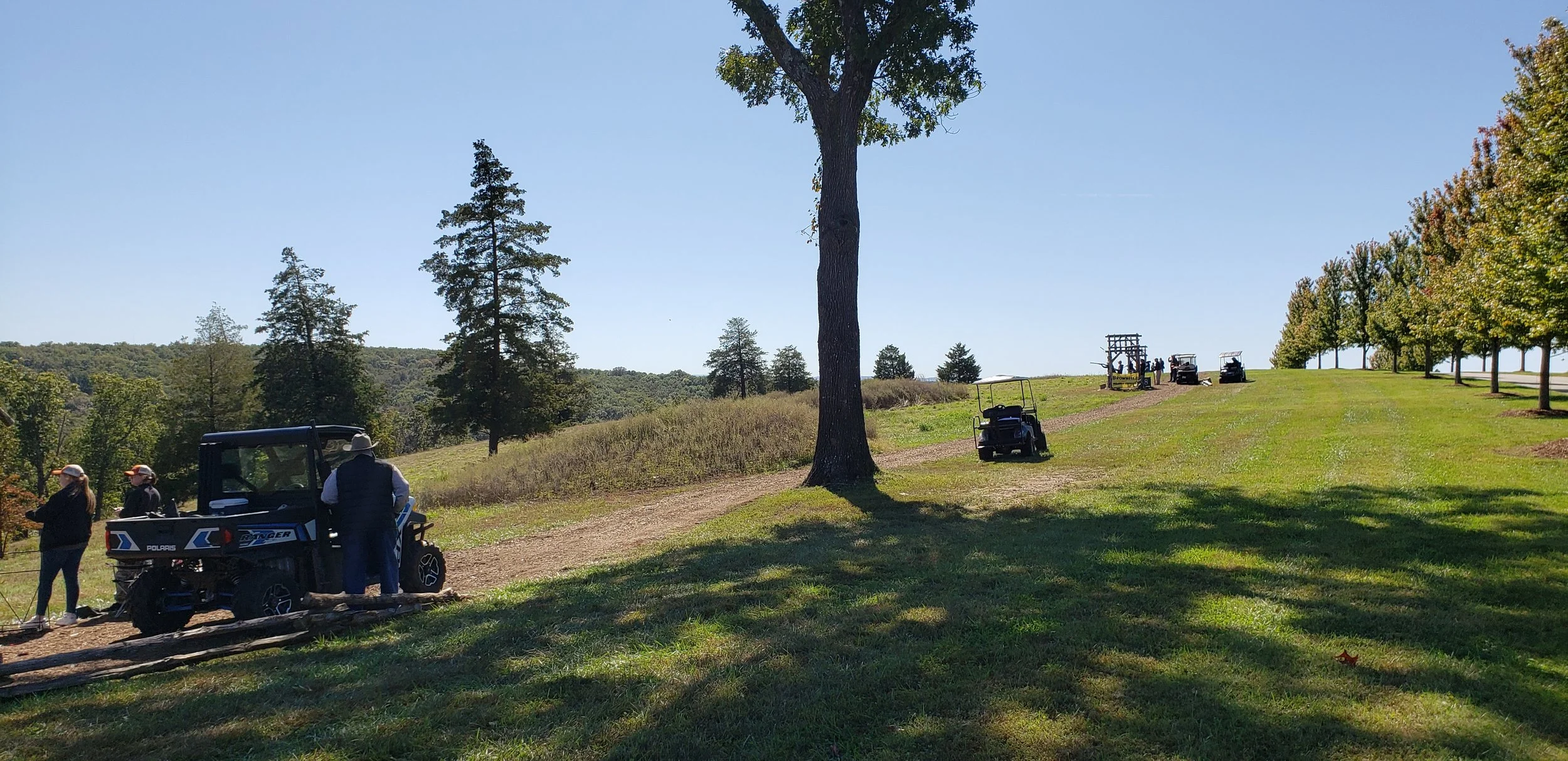 People standing next to golf carts on a golf course with a line of golf carts in the distance under a clear blue sky, surrounded by trees and grassy hills.