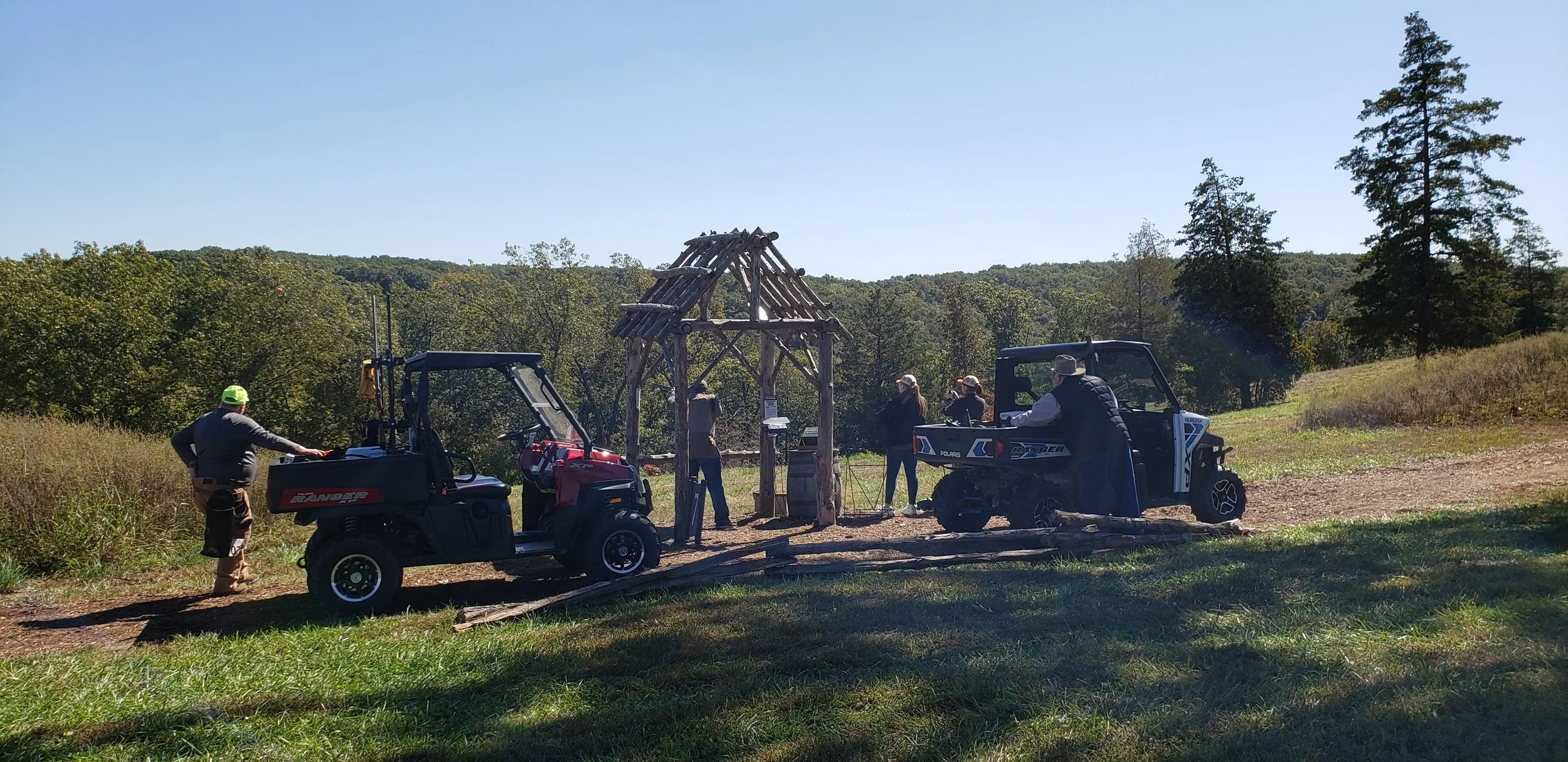 Group of people outdoors near wooden structure with two utility vehicles on grassy trail, surrounded by trees and hills, sunny day.