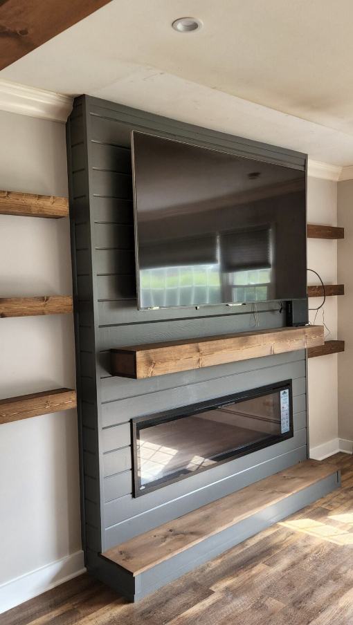 Living room fireplace with mounted flat-screen TV, gray shiplap wall, wooden mantel, and floating wooden shelves on either side.