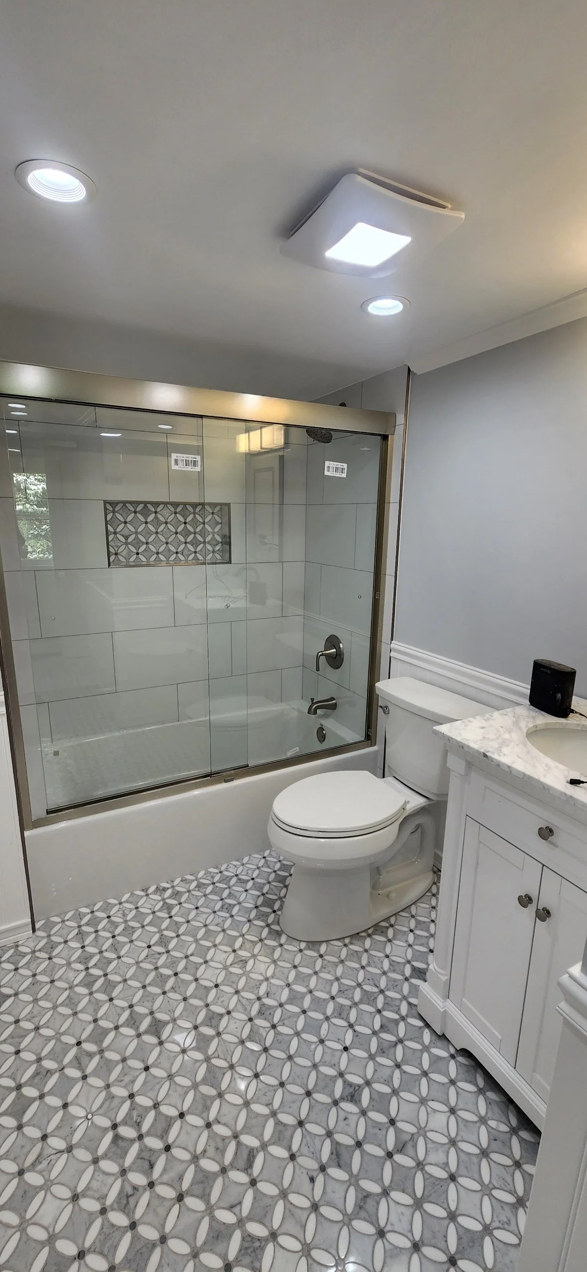 Bathroom with a white toilet, a white vanity with a marble countertop, a glass shower enclosure with patterned tiles, and patterned floor tiles.