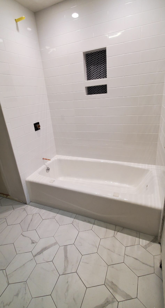 Empty bathroom with white tile walls and hexagon marble-patterned floor, showing bathtub, wall vent, and plumbing fixtures.