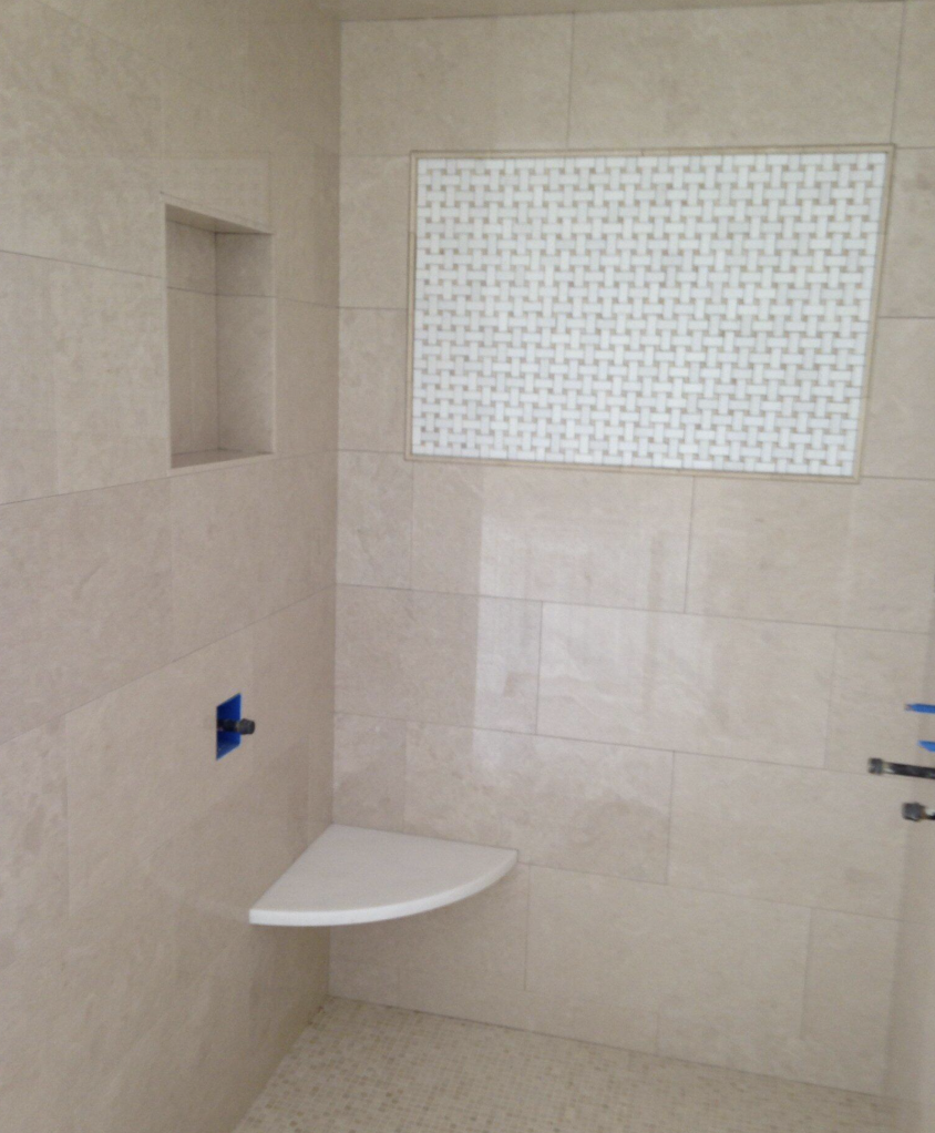 A shower space with beige tiles, a small built-in shelf, and a corner seat. There is a window with a woven pattern covering and blue tape on the wall.