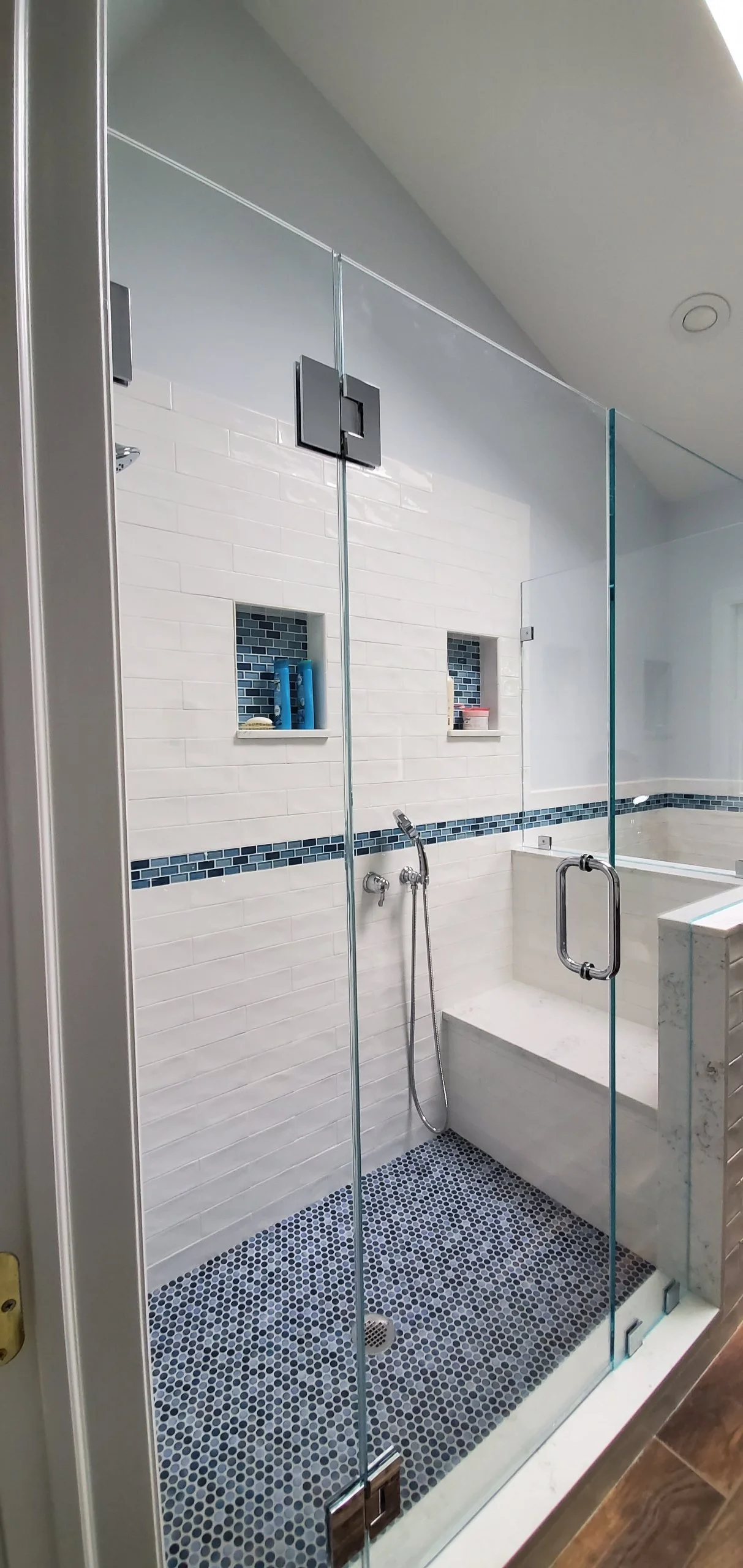 A modern bathroom shower with white subway tile walls, small black and white mosaic floor tiles, and built-in niche shelves holding toiletries. The shower is enclosed with clear glass doors and features a wall-mounted shower head and hand-held shower wand.
