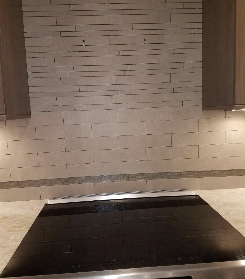 Kitchen stove with a black glass cooktop, stainless steel trim, and beige marble countertops in front of a tiled wall with horizontal rectangular tiles.