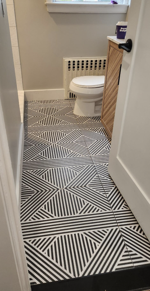 View of a bathroom with a geometric black and white patterned tile floor, a white toilet, a wooden cabinet, and a window with a small purple decoration.