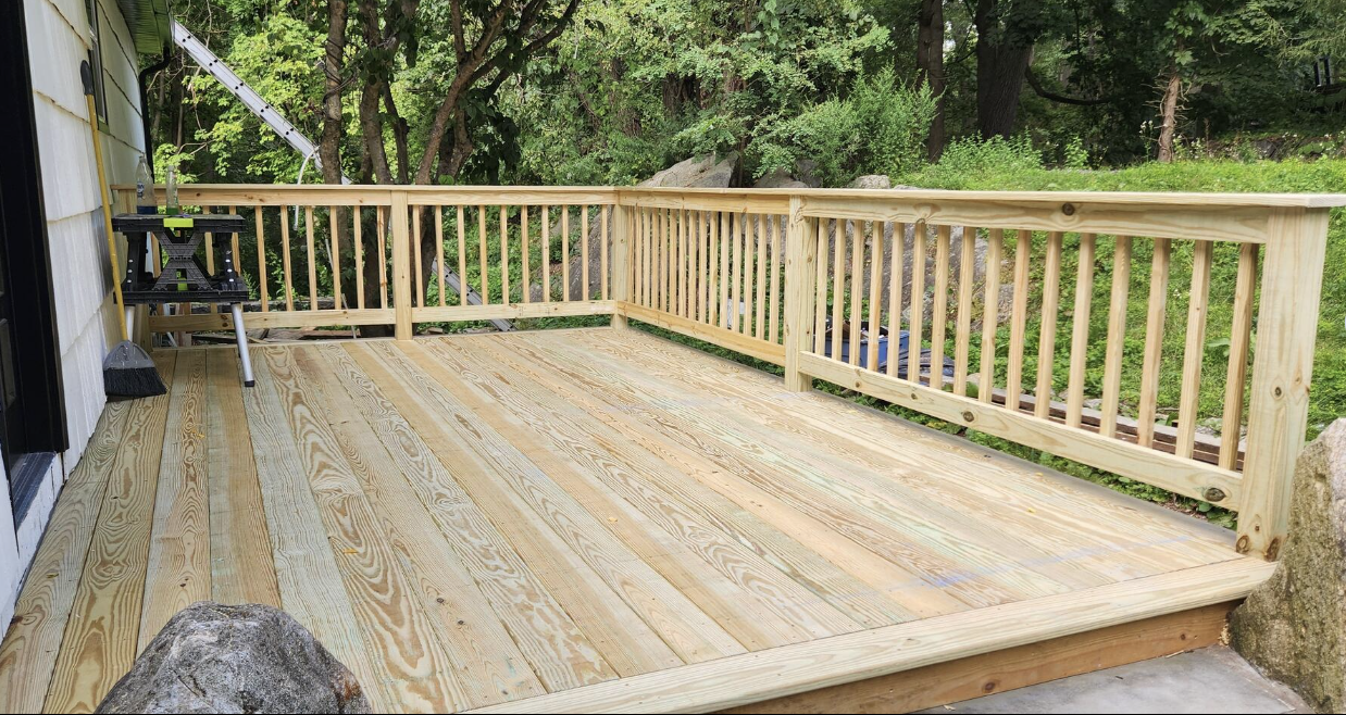 Newly built wooden deck with railing, located outside a house with tools and construction materials nearby, surrounded by trees and greenery.