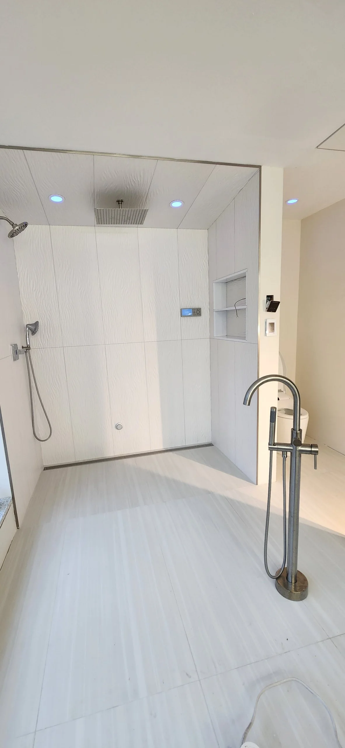 Empty modern shower with white tiled walls and ceiling, two showerheads, a built-in niche, a digital control panel, and a metal handrail, with ceiling recessed lights.