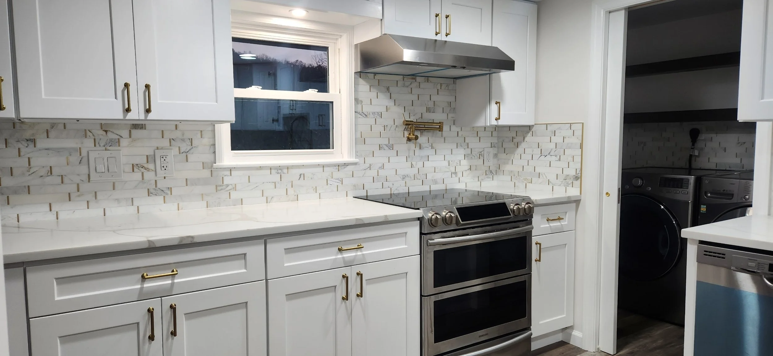 Modern white kitchen with marble countertops, white cabinets with gold handles, stainless steel oven and range hood, white subway tile backsplash, window above the sink, and laundry washer and dryer in the adjacent room.