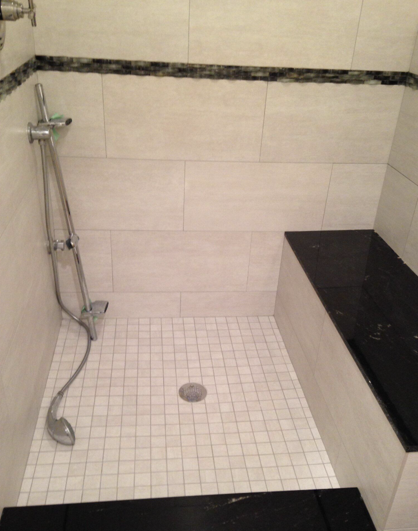 Shower area with handheld showerhead, tiled walls, small tiled floor, and built-in bench with black surface.