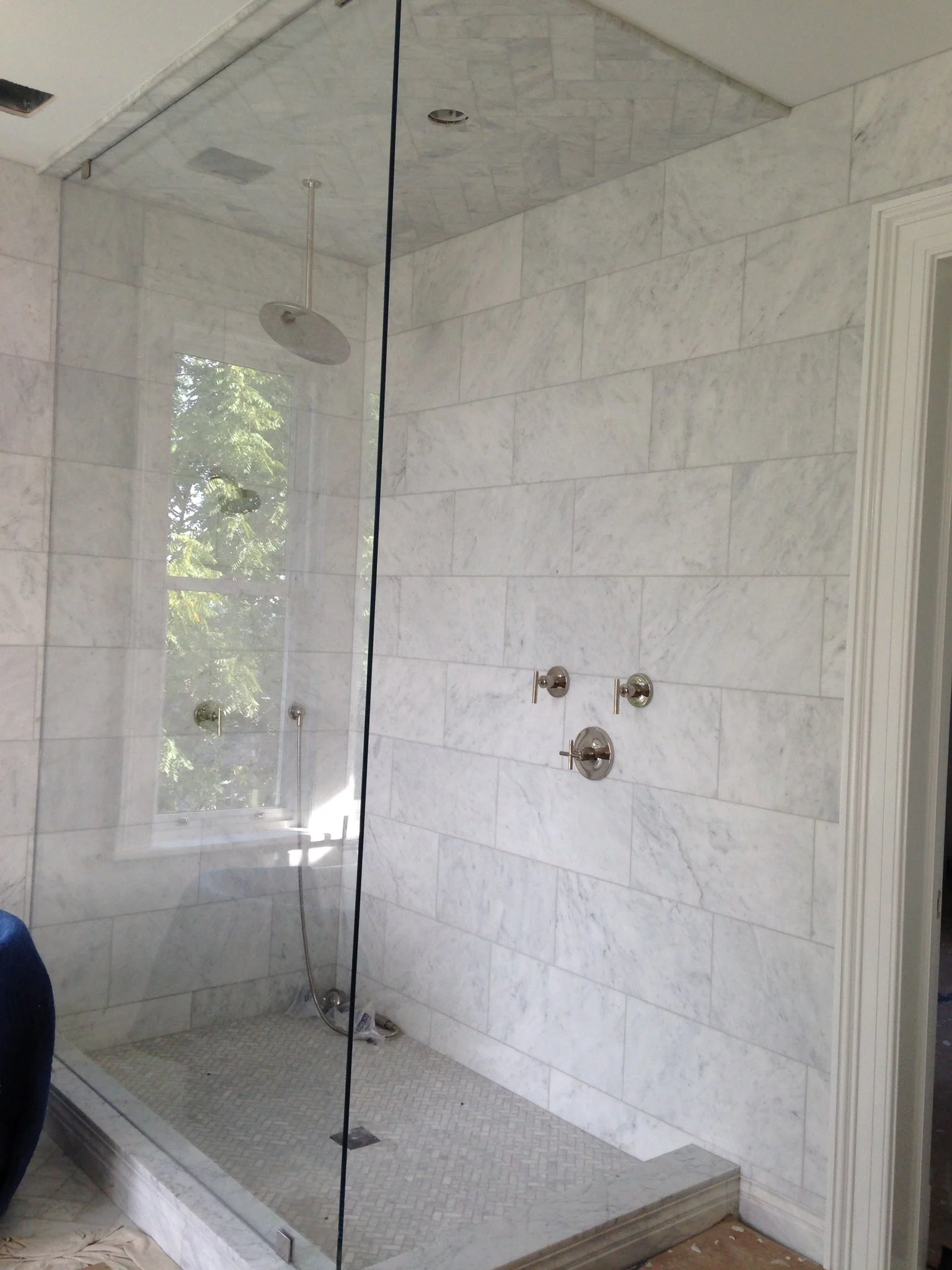 A modern walk-in shower with white marble tiles, a glass partition, a rainfall showerhead, and multiple control knobs.