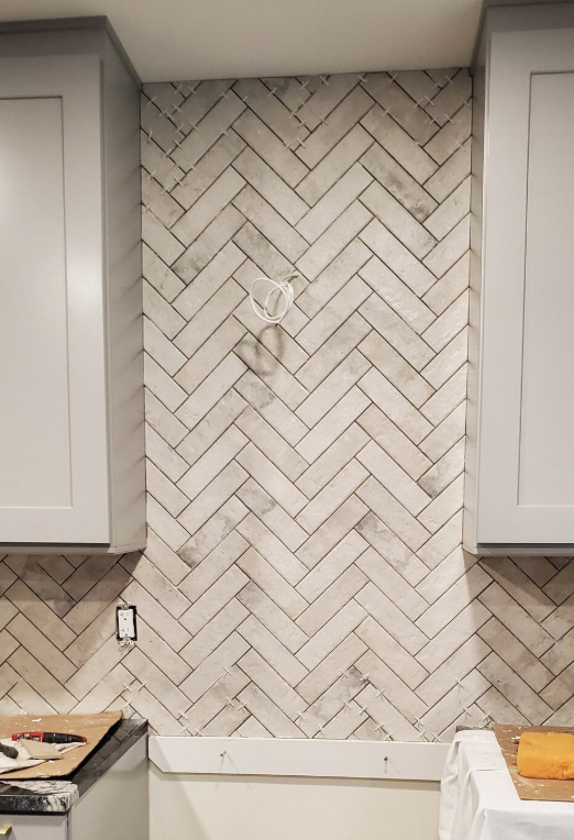 New herringbone-style tile backsplash being installed in a kitchen between two light gray cabinets, with a white countertop below and some tools on the counter.