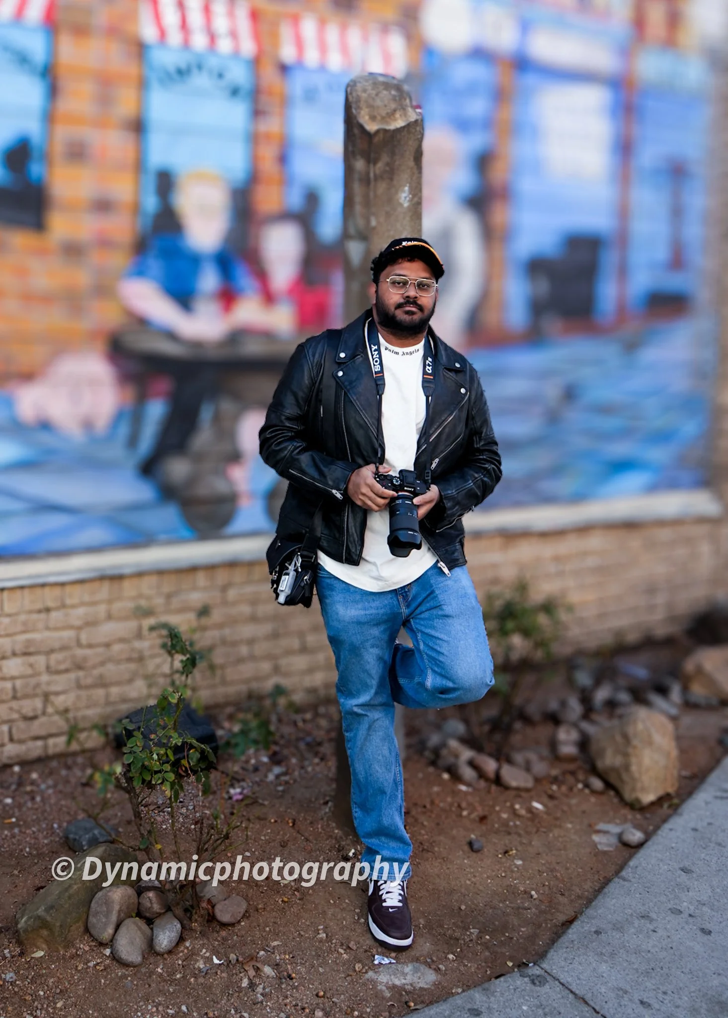 A man holding a camera, standing with one leg propped against a small patch of ground with rocks and a small bush, in front of a colorful mural on a brick wall.