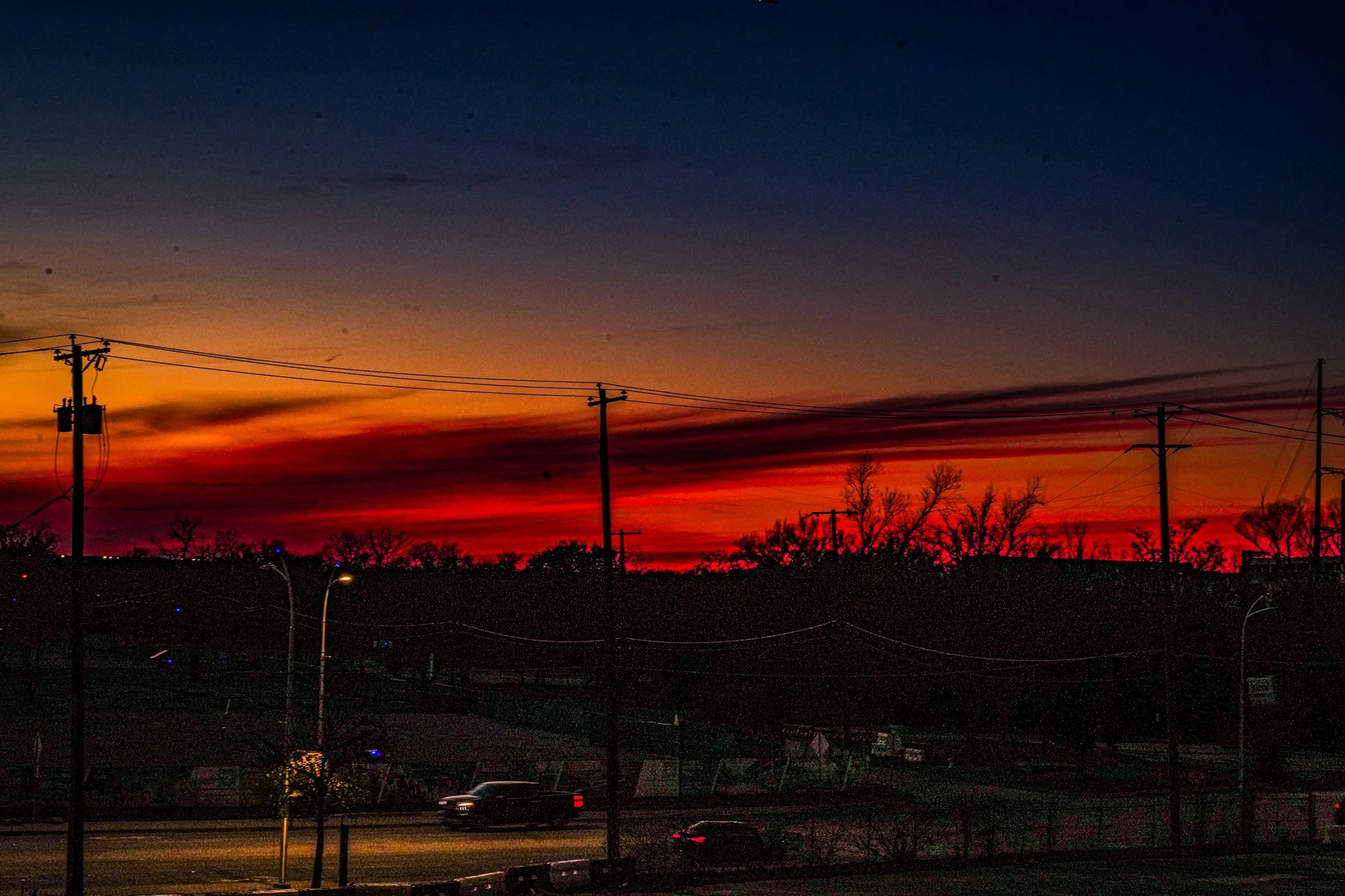 Dusk sky with vibrant orange, red, purple, and dark blue hues over silhouetted trees, power lines, and a road with moving cars.